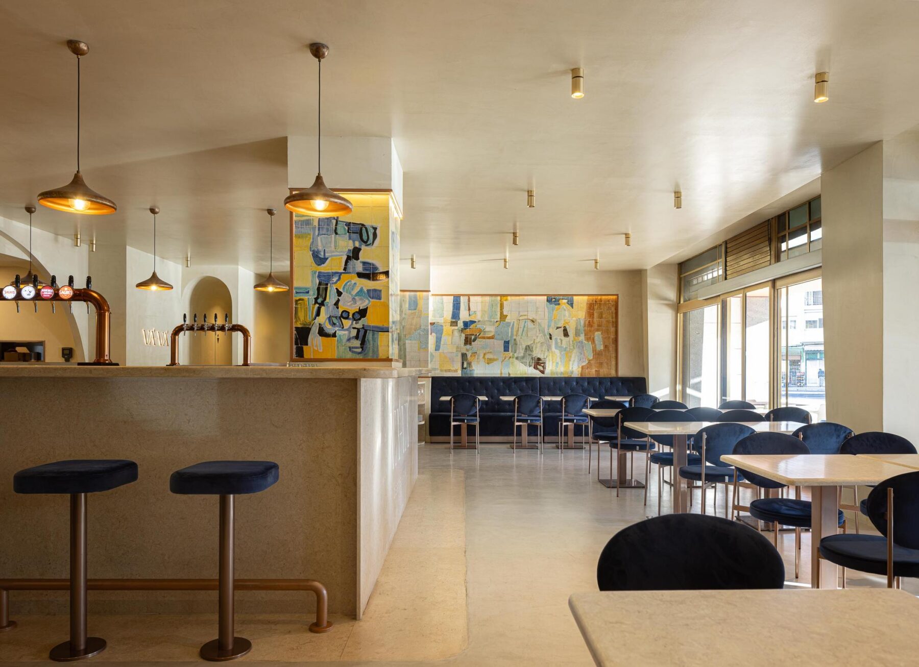 Salão: Balcão em pedra clara com bancos altos de veludo azul, pavimento em cimento afagado e candeeiros suspensos em metal. — Cervejaria Vává - Requalificação arquitetura Rethorica Studio em Lisboa