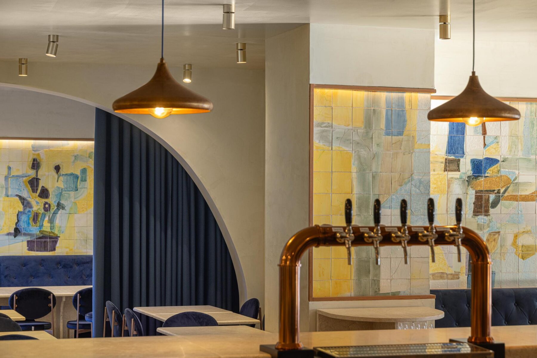 Salão: Arco divide o espaço, com cortina azul escura, balcão, painel de azulejos e candeeiros suspensos. — Cervejaria Vává - Requalificação arquitetura Rethorica Studio em Lisboa