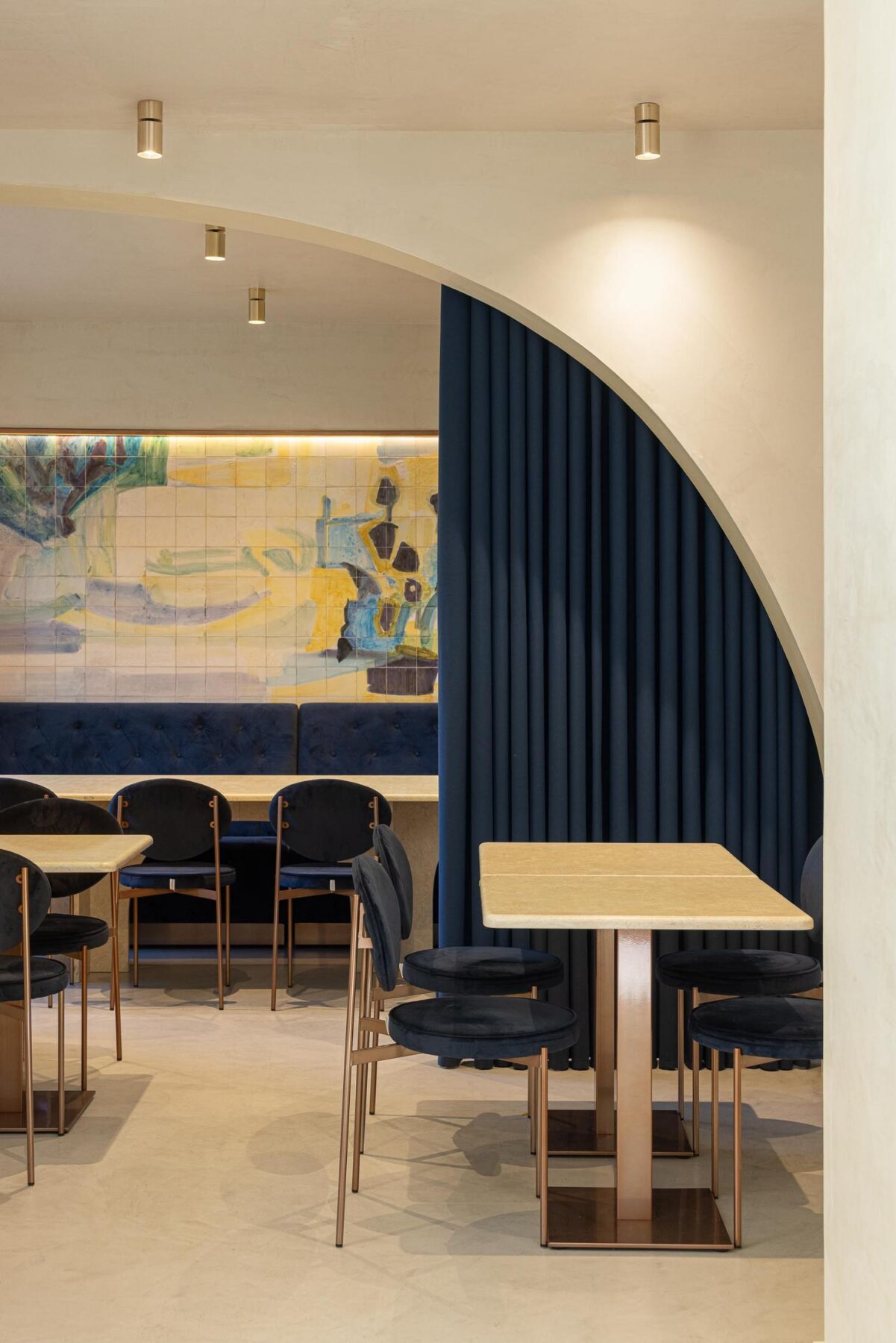 Salão: Mesa com cadeiras de veludo azul e estrutura metálica, separada por um arco com cortina azul. — Cervejaria Vává - Requalificação arquitetura Rethorica Studio em Lisboa