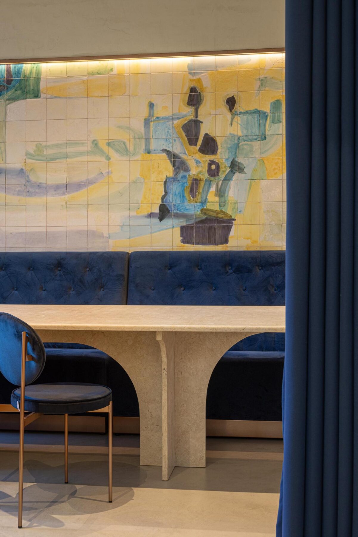 Salão: Mesa com tampo em pedra clara e cadeiras de veludo azul, com painel de azulejos ao fundo. — Cervejaria Vává - Requalificação arquitetura Rethorica Studio em Lisboa