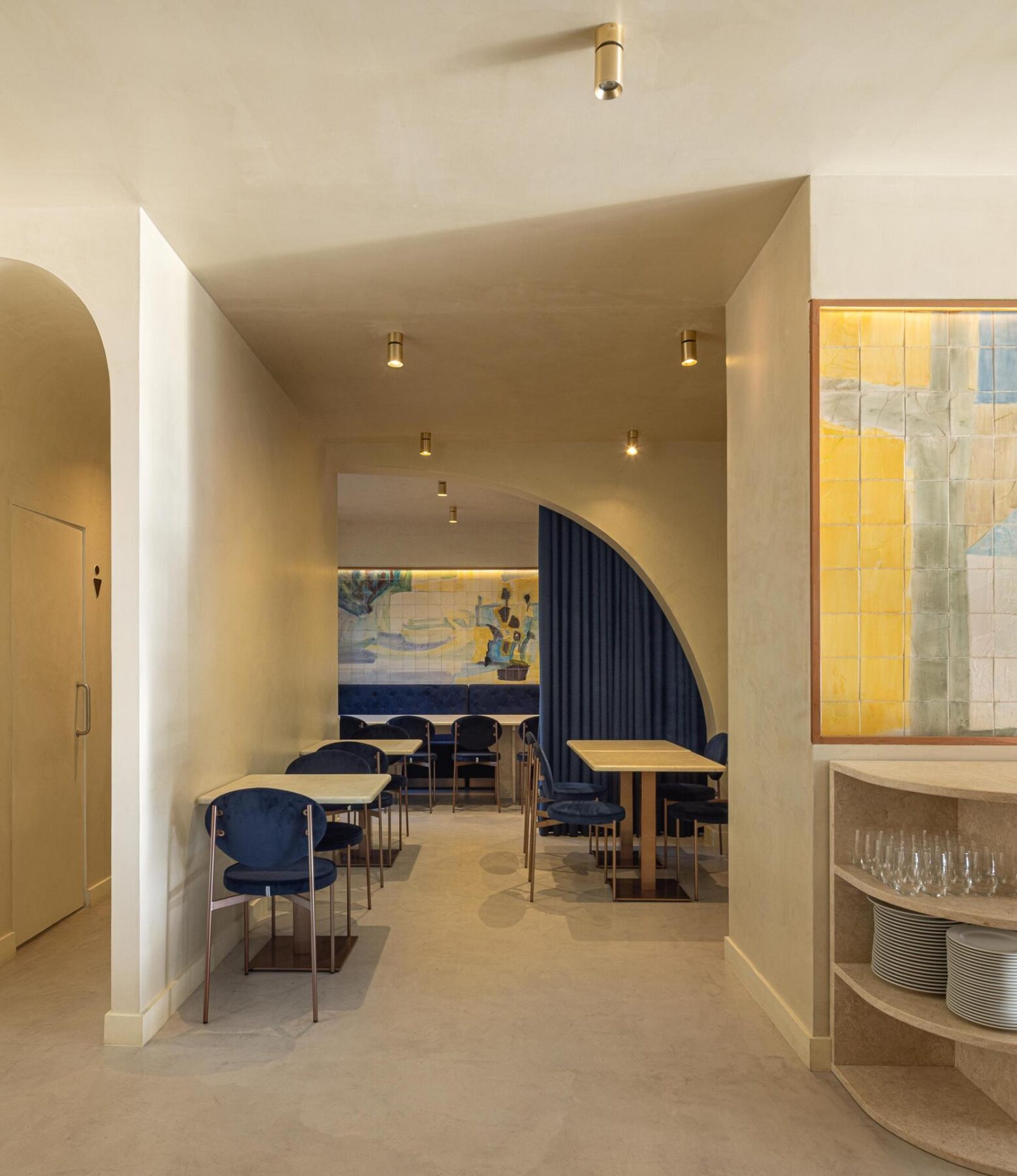Salão: Corredor com arcos, mesas com cadeiras de veludo azul, painel de azulejos e prateleira com louça. — Cervejaria Vává - Requalificação arquitetura Rethorica Studio em Lisboa