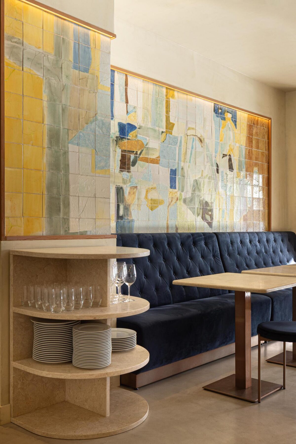 Salão: Sofá corrido de veludo azul, mesa de mármore, prateleira com louça e painel de azulejos. — Cervejaria Vává - Requalificação arquitetura Rethorica Studio em Lisboa