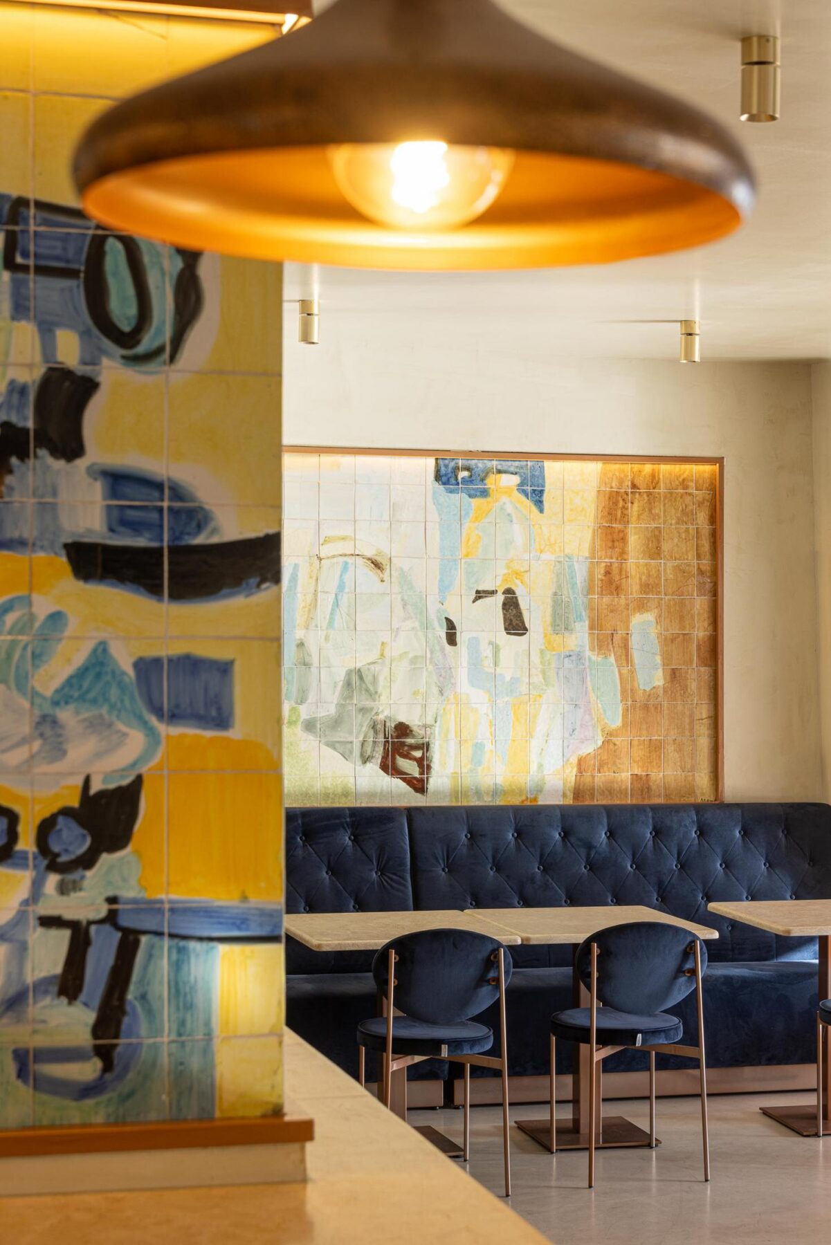 Salão: Candeeiro suspenso, painel de azulejos, sofá corrido de veludo azul e mesas com cadeiras. — Cervejaria Vává - Requalificação arquitetura Rethorica Studio em Lisboa