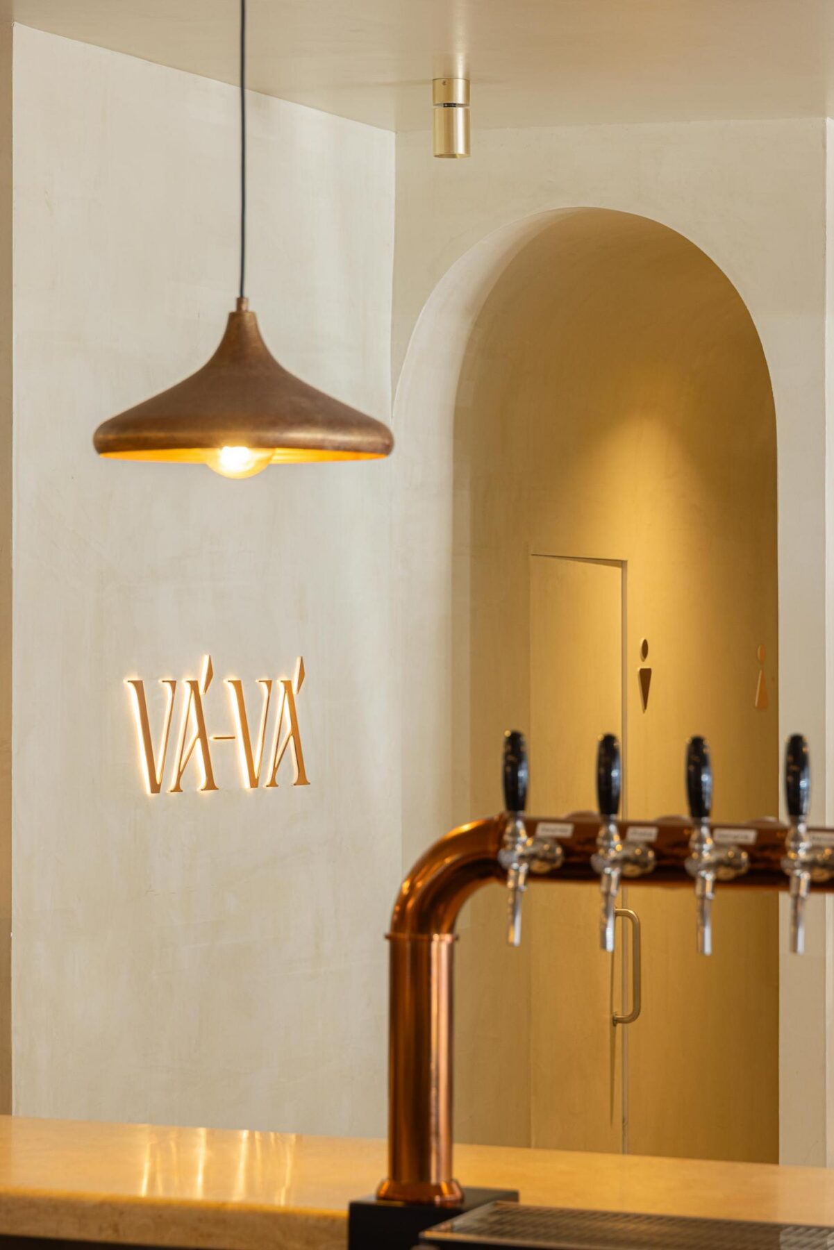 Balcão: Torneiras de cerveja em cobre, candeeiro suspenso e sinalética "Vá-Vá" na parede. — Cervejaria Vává - Requalificação arquitetura Rethorica Studio em Lisboa