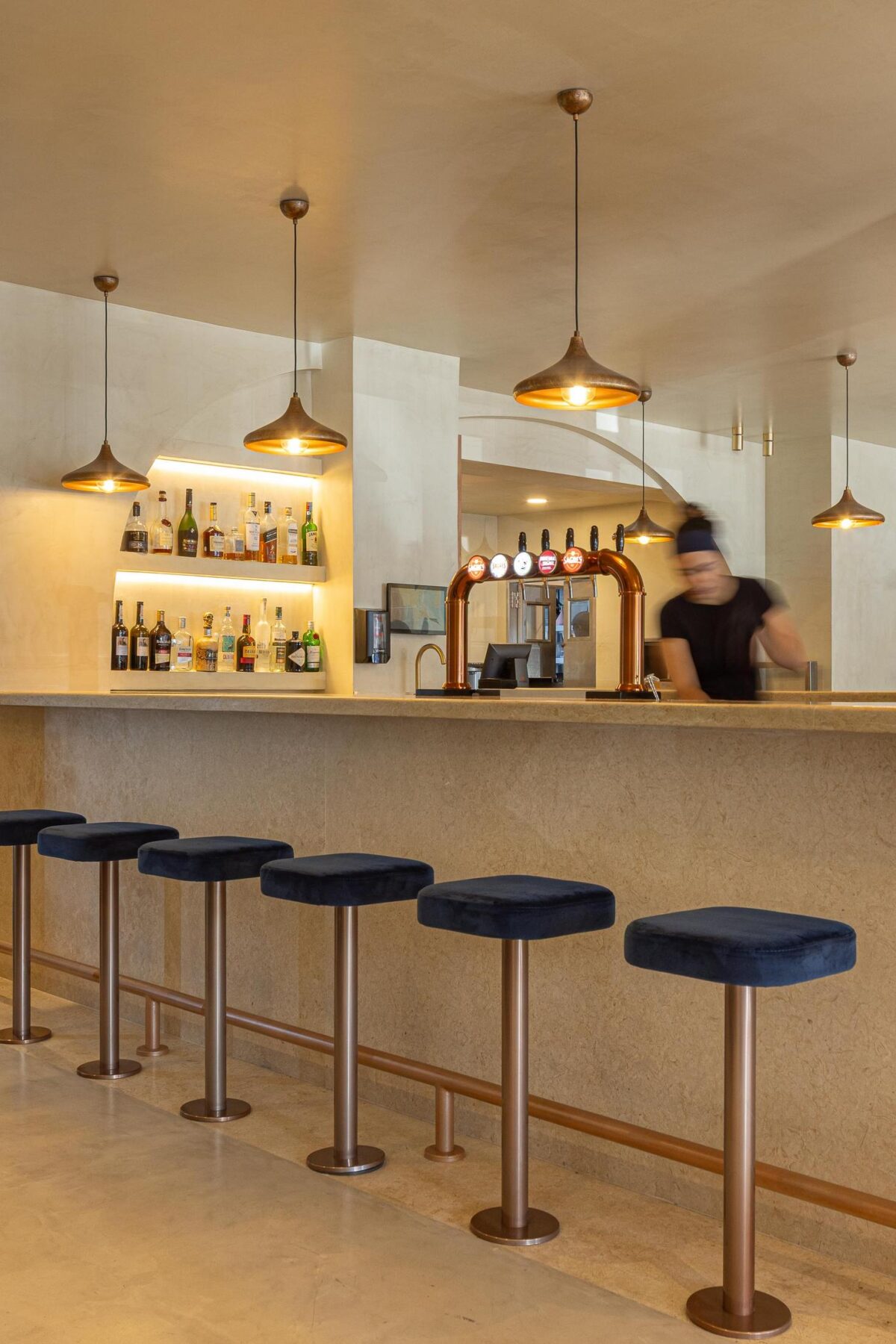 Balcão: Balcão com bancos altos de veludo azul, garrafeira iluminada e candeeiros suspensos. — Cervejaria Vává - Requalificação arquitetura Rethorica Studio em Lisboa