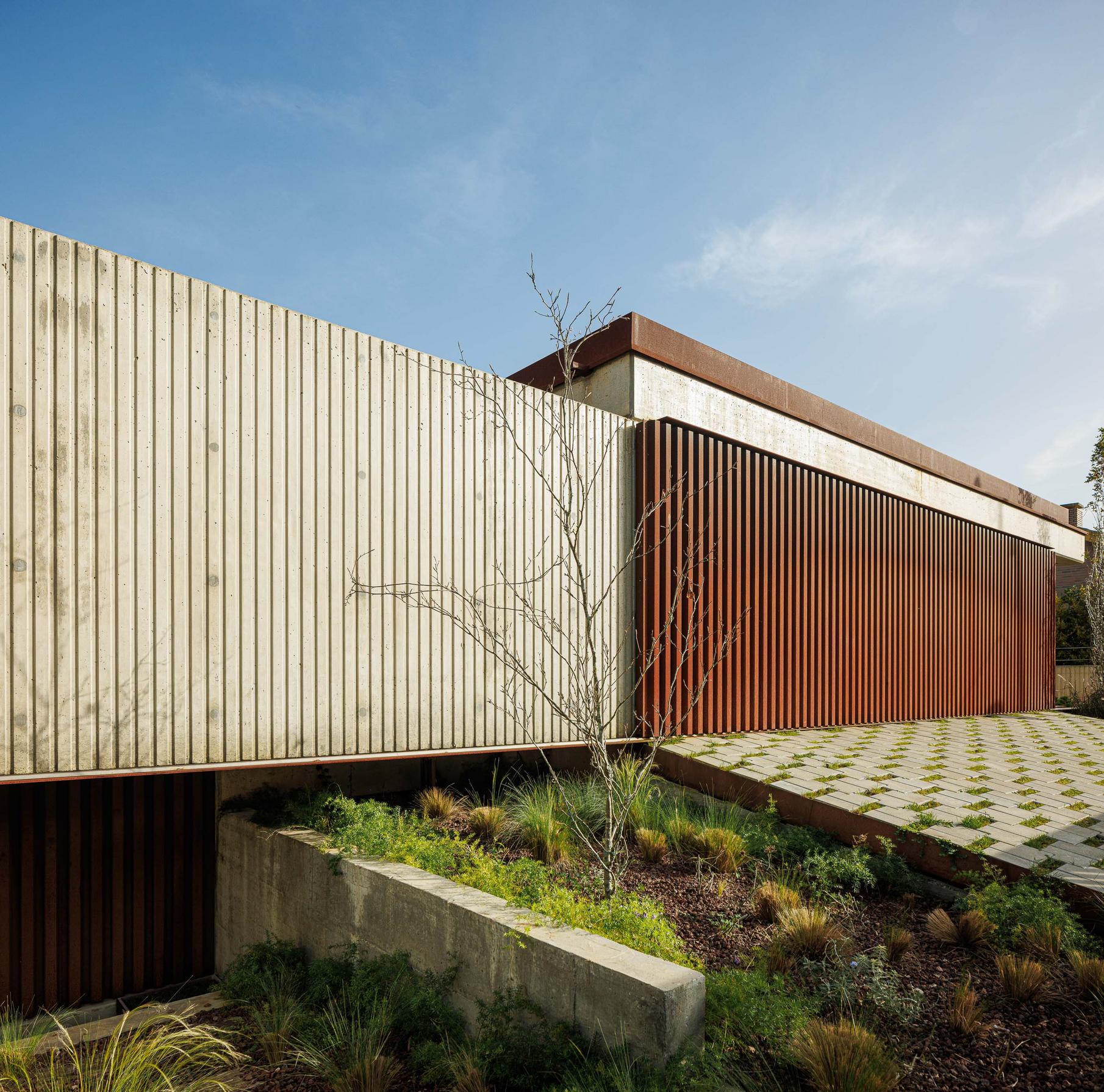 Detalhe da fachada com ripas verticais de madeira e betão aparente, jardim com vegetação rasteira e pavimento em grelha. — Mimosa House arquitetura Pedro Henrique Arquitecto em Esmoriz