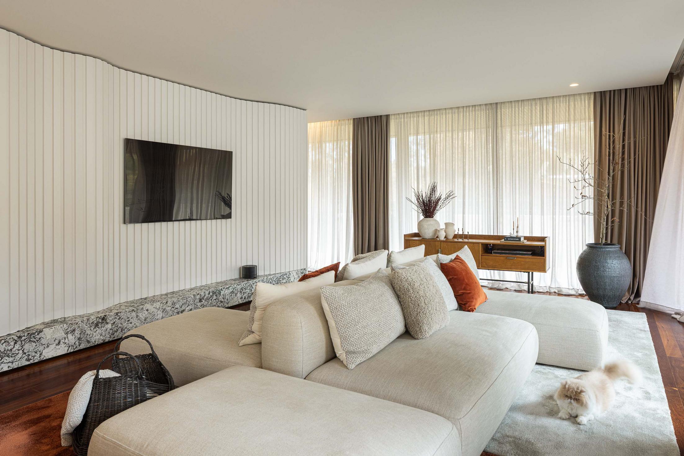 Sala de estar com sofá modular, alcatifa clara, cortinas e parede revestida a ripas brancas. — Mimosa House arquitetura Pedro Henrique Arquitecto em Esmoriz