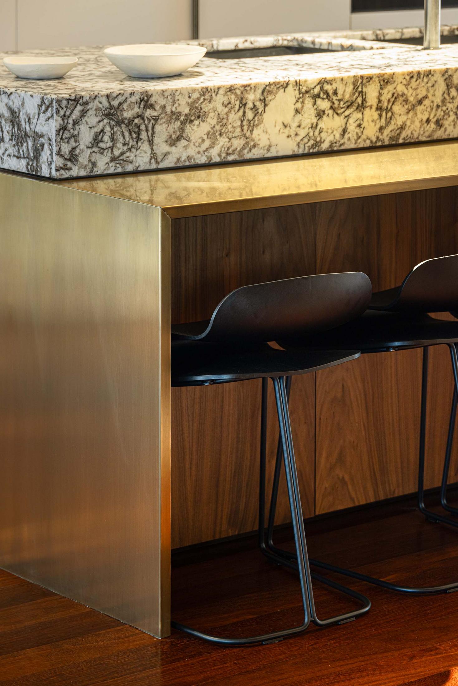 Detalhe da ilha da cozinha com bancada em mármore, revestimento em latão e cadeiras pretas. — Mimosa House arquitetura Pedro Henrique Arquitecto em Esmoriz