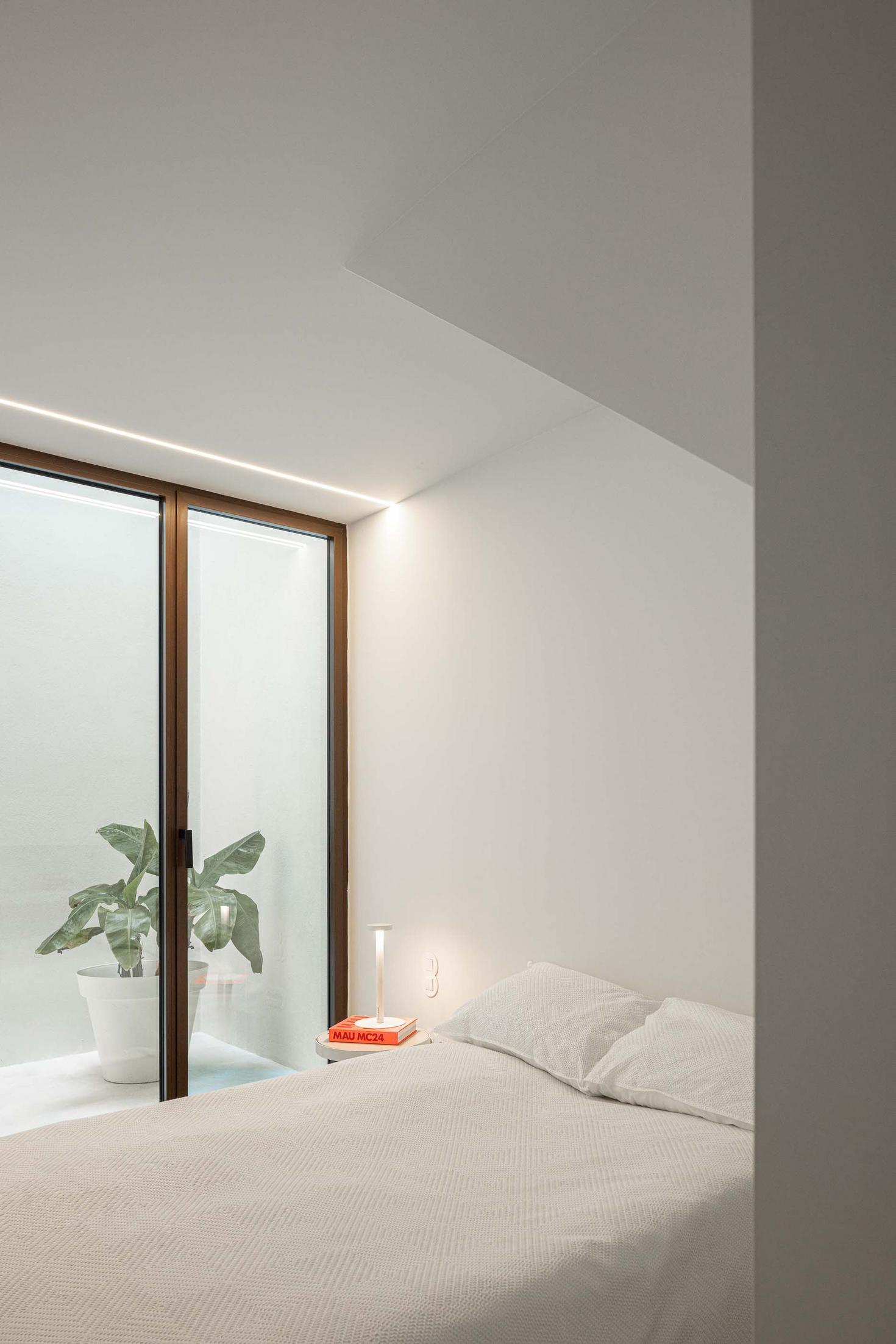 Quarto com cama branca e candeeiro de mesa. Vão envidraçado vertical com planta, proporcionando iluminação zenital. Paredes brancas — Beira Mar II arquitectura Paulo Martins fotografia Ivo Tavares