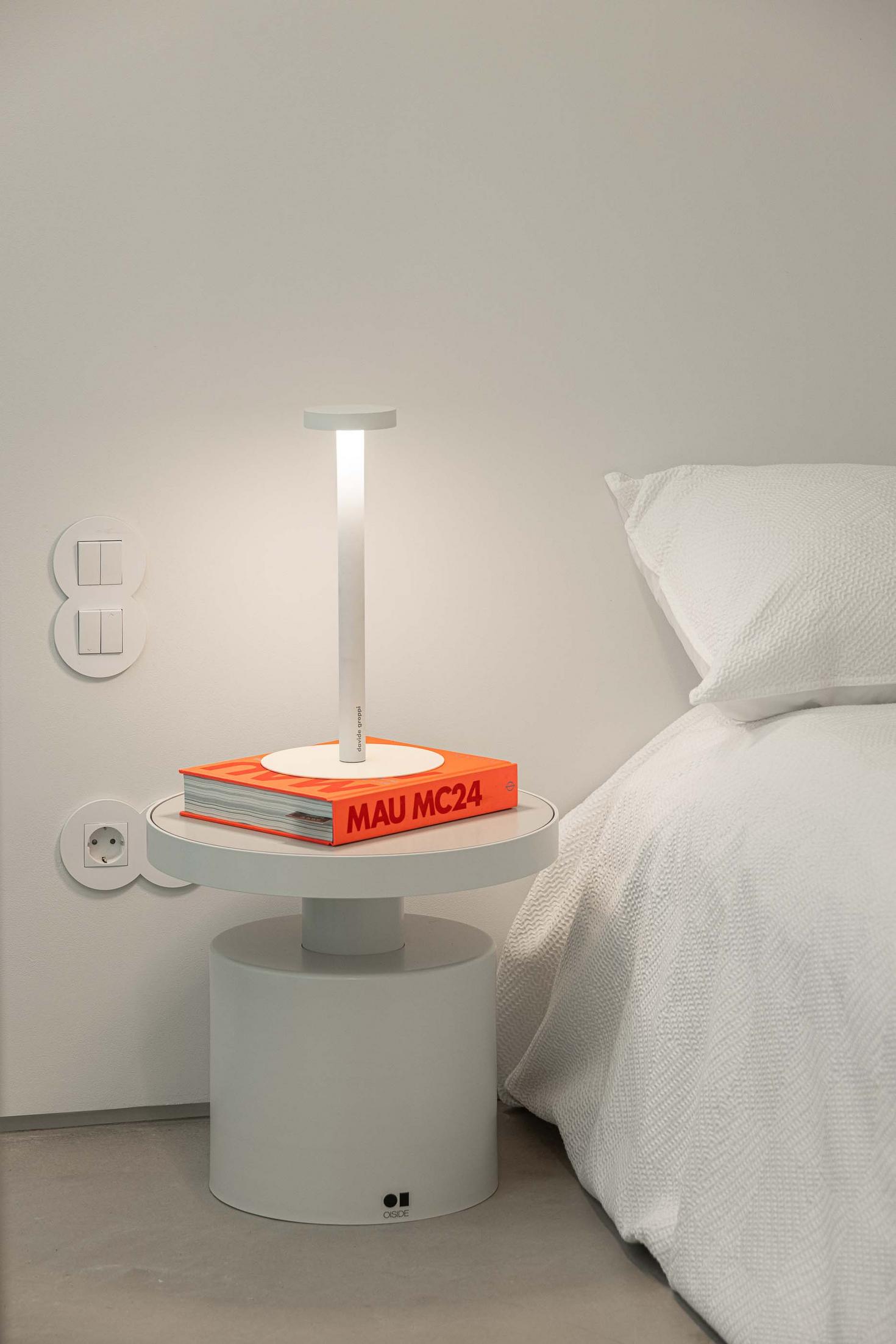 Detalhe de mesa de cabeceira branca com candeeiro de mesa e livro. Cama com roupa de cama branca. Tomadas e interruptores brancos — Beira Mar II arquitectura Paulo Martins fotografia Ivo Tavares