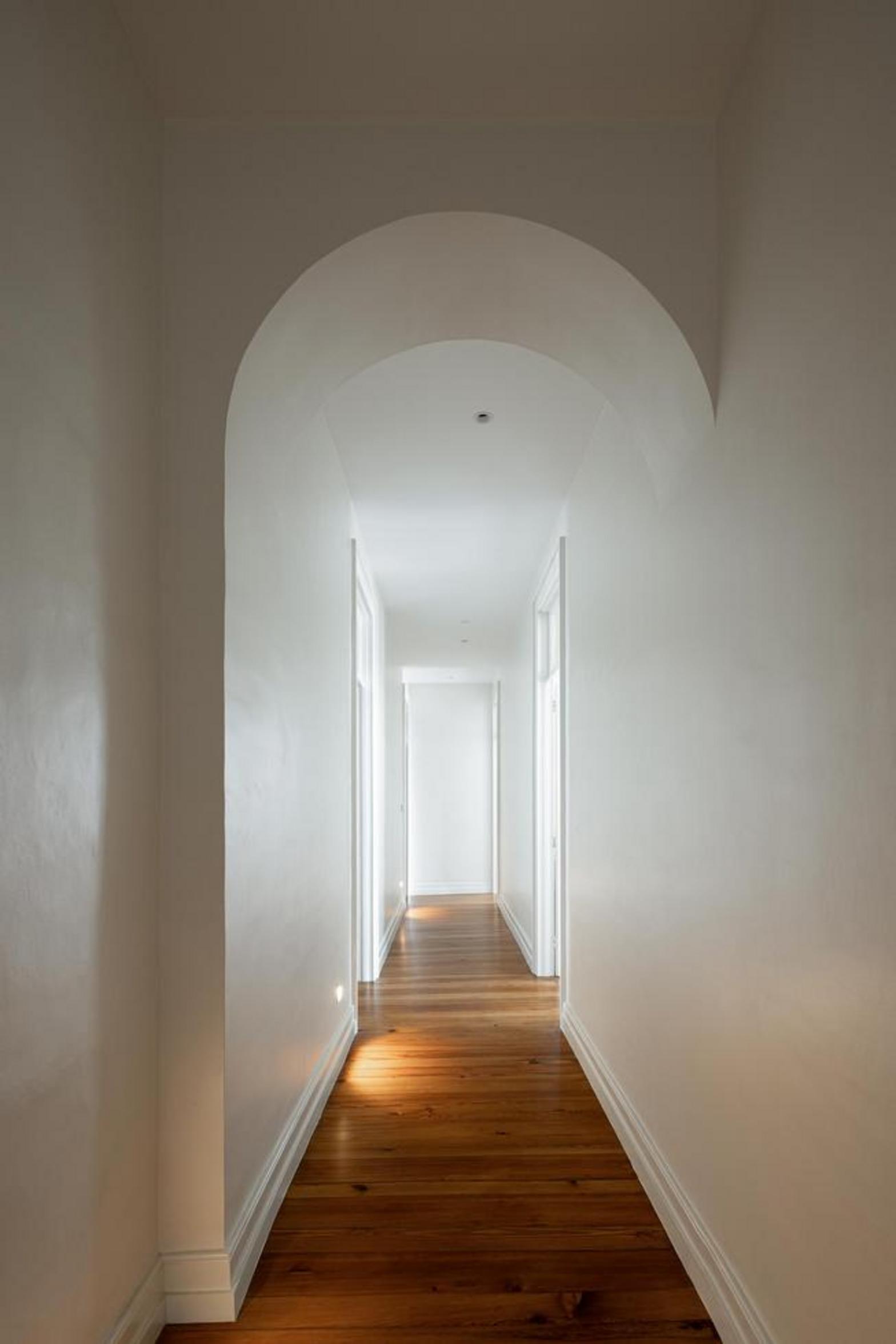 Corredor. Pavimento em madeira mel, paredes brancas. Vários arcos de porta. Homem de costas, corrimão branco — Casa T+A Açores arquitectura Lopes da Costa fotografia Ivo Tavares