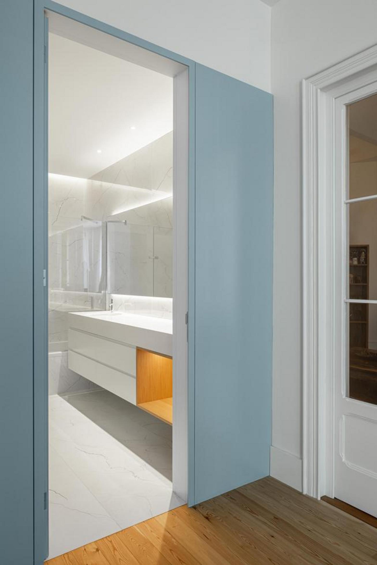 Interior. Pavimento em madeira mel, paredes brancas. Painel azul claro com porta semi-aberta revelando casa de banho — Casa T+A Açores arquitectura Lopes da Costa fotografia Ivo Tavares