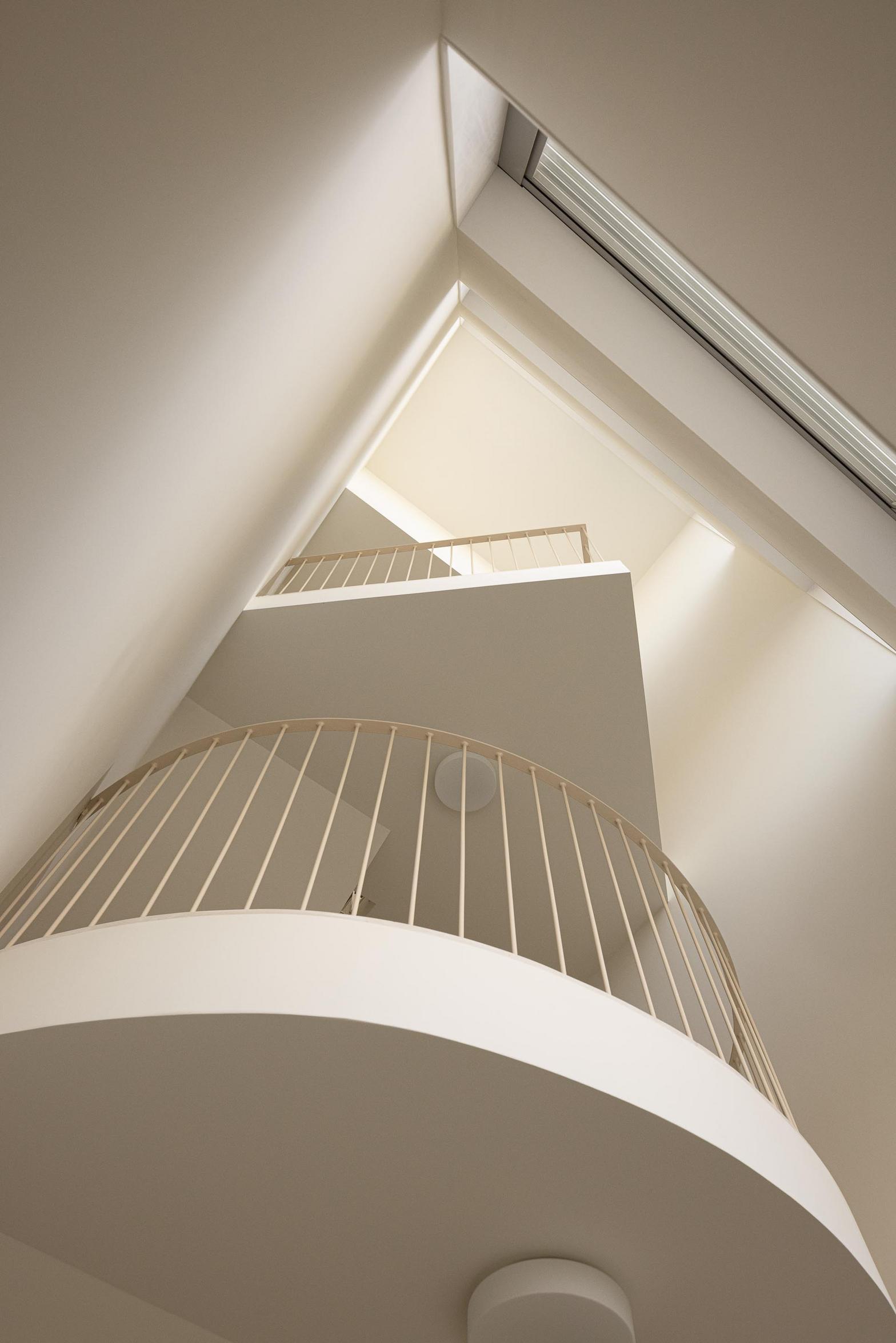 Escadaria interior com paredes brancas e guardas metálicas claras. A luz zenital acentua a volumetria curva e a verticalidade do espaço — Edifício Multifamiliar Hintze Ribeiro arquitectura Helder da Rocha fotografia Ivo Tavares