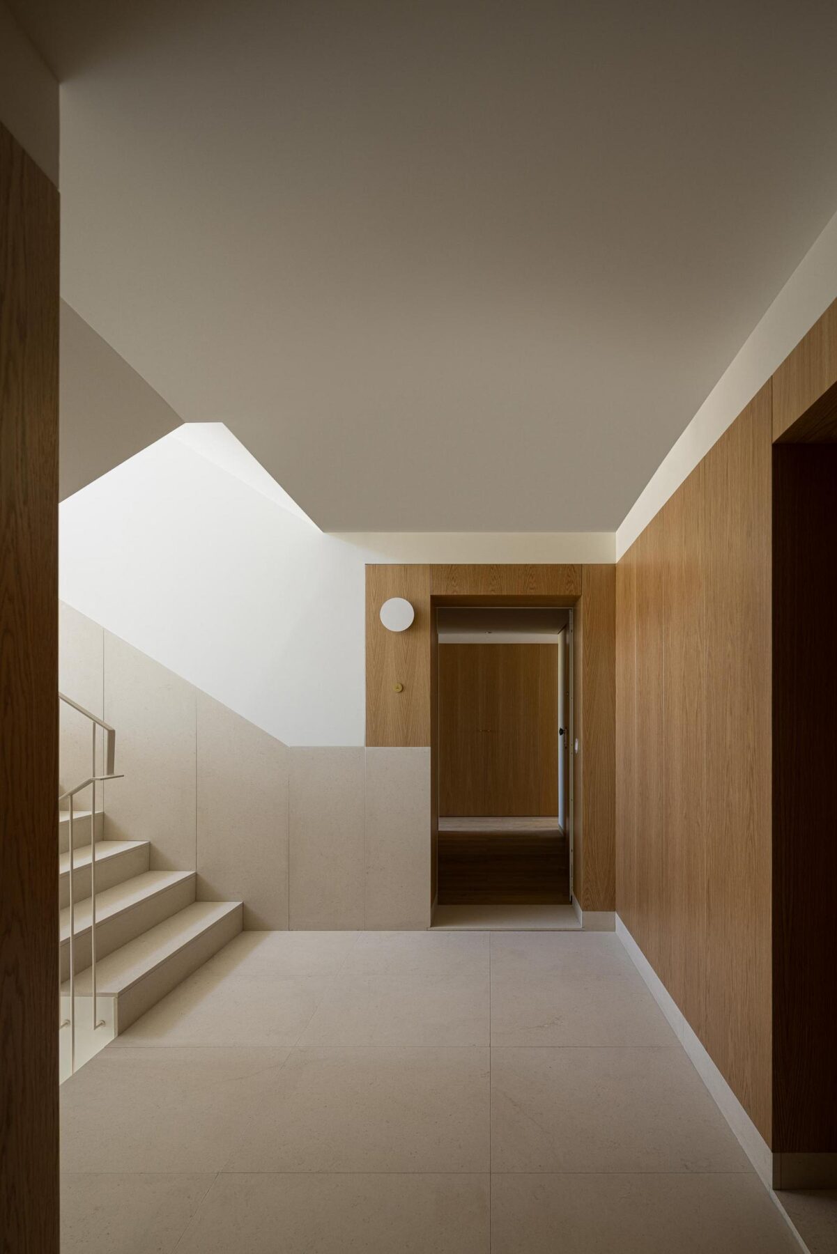 Hall de entrada com escada em pedra clara, revestimento em madeira de carvalho e iluminação zenital, criando um ambiente acolhedor. — EDIFÍCIO DE HABITAÇÃO MULTIFAMILIAR HINTZE RIBEIRO arquitectura HELDER DA ROCHA ARQUITECTOS fotografia Ivo Tavares