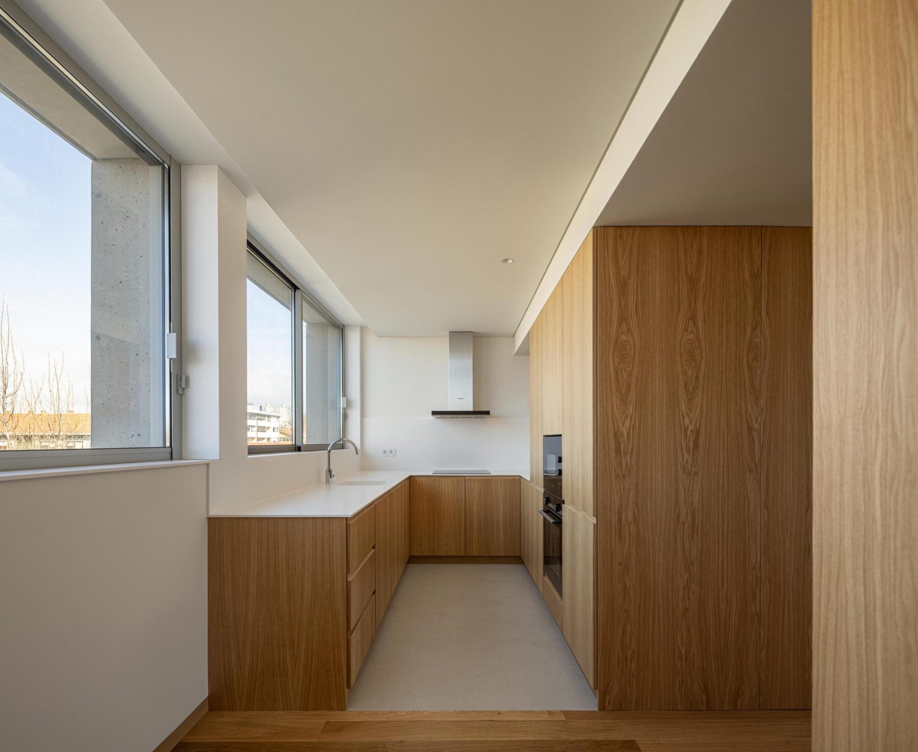 Cozinha com armários em madeira de carvalho, bancada branca e pavimento em microcimento cinzento, com amplas janelas para o exterior. — EDIFÍCIO DE HABITAÇÃO MULTIFAMILIAR HINTZE RIBEIRO arquitectura HELDER DA ROCHA ARQUITECTOS fotografia Ivo Tavares