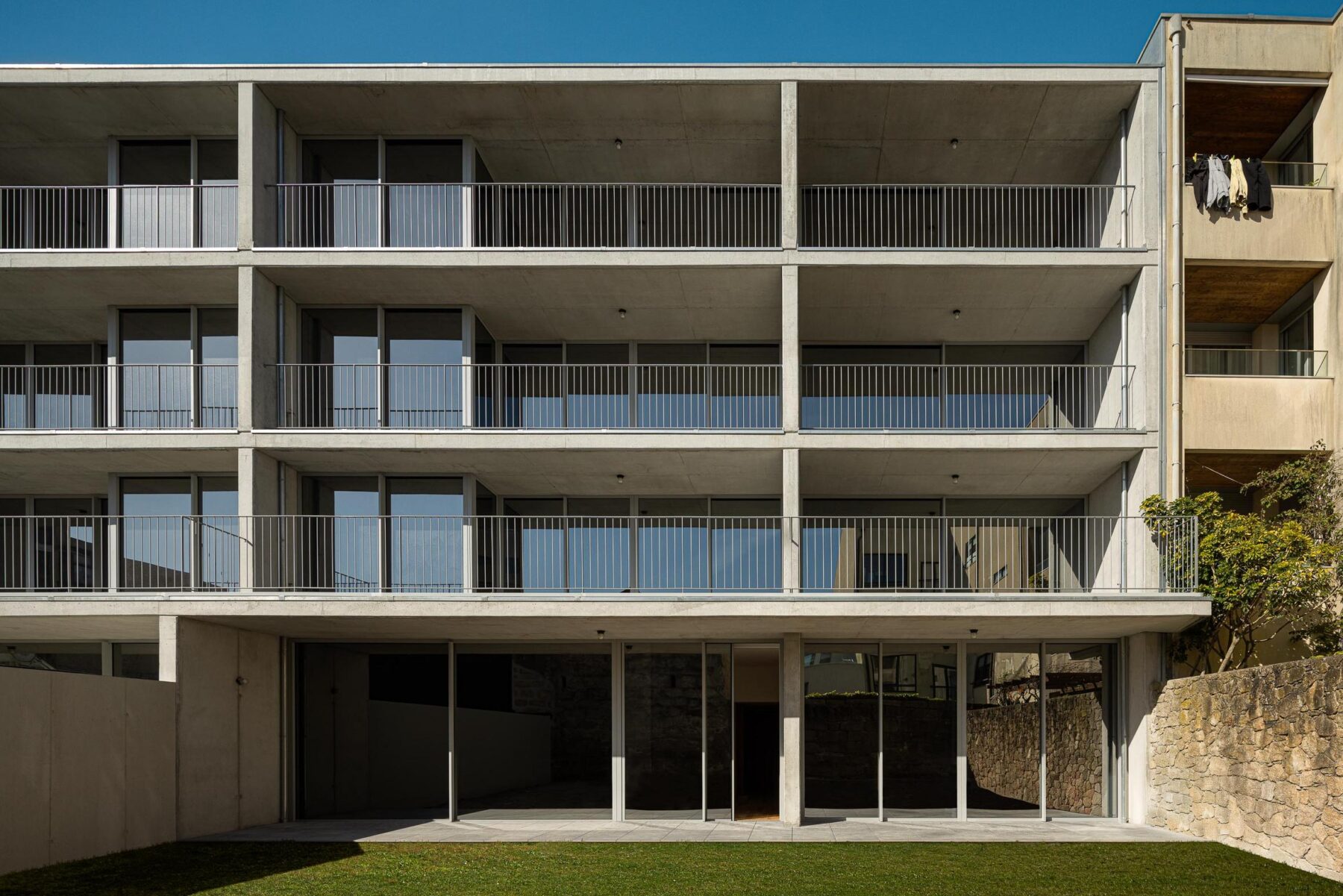 Fachada posterior do edifício com varandas em betão aparente e jardim relvado, evidenciando a relação entre interior e exterior. — EDIFÍCIO DE HABITAÇÃO MULTIFAMILIAR HINTZE RIBEIRO arquitectura HELDER DA ROCHA ARQUITECTOS fotografia Ivo Tavares