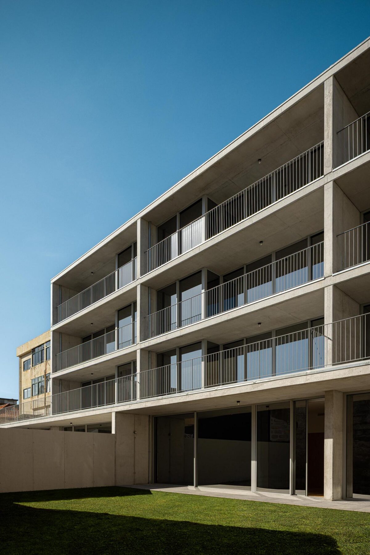 Fachada posterior do edifício com varandas em betão aparente e jardim relvado, integrando o edifício no contexto urbano e natural. — EDIFÍCIO DE HABITAÇÃO MULTIFAMILIAR HINTZE RIBEIRO arquitectura HELDER DA ROCHA ARQUITECTOS fotografia Ivo Tavares