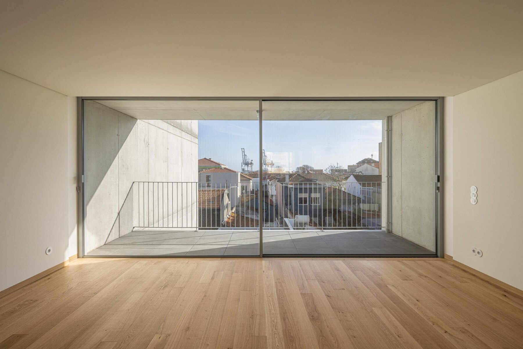 Sala ampla com pavimento em madeira de carvalho e varanda com guarda metálica, com vista para a paisagem urbana de Leça da Palmeira. — EDIFÍCIO DE HABITAÇÃO MULTIFAMILIAR HINTZE RIBEIRO arquitectura HELDER DA ROCHA ARQUITECTOS fotografia Ivo Tavares