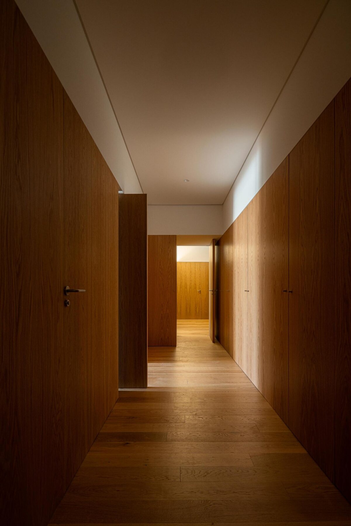 Corredor interior com pavimento e revestimento em madeira de carvalho, portas embutidas e iluminação linear no teto branco. — EDIFÍCIO DE HABITAÇÃO MULTIFAMILIAR HINTZE RIBEIRO arquitectura HELDER DA ROCHA ARQUITECTOS fotografia Ivo Tavares
