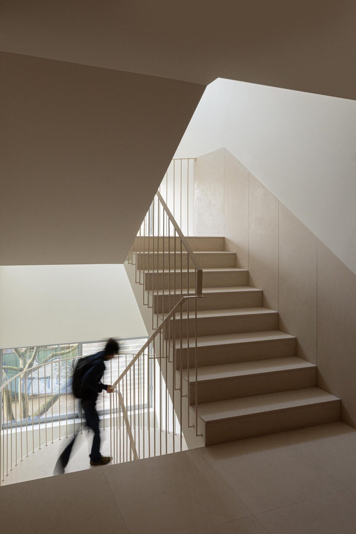 Escada em pedra clara com guarda metálica, iluminada zenitalmente, com um utilizador a subir, destacando a verticalidade do espaço. — EDIFÍCIO DE HABITAÇÃO MULTIFAMILIAR HINTZE RIBEIRO arquitectura HELDER DA ROCHA ARQUITECTOS fotografia Ivo Tavares