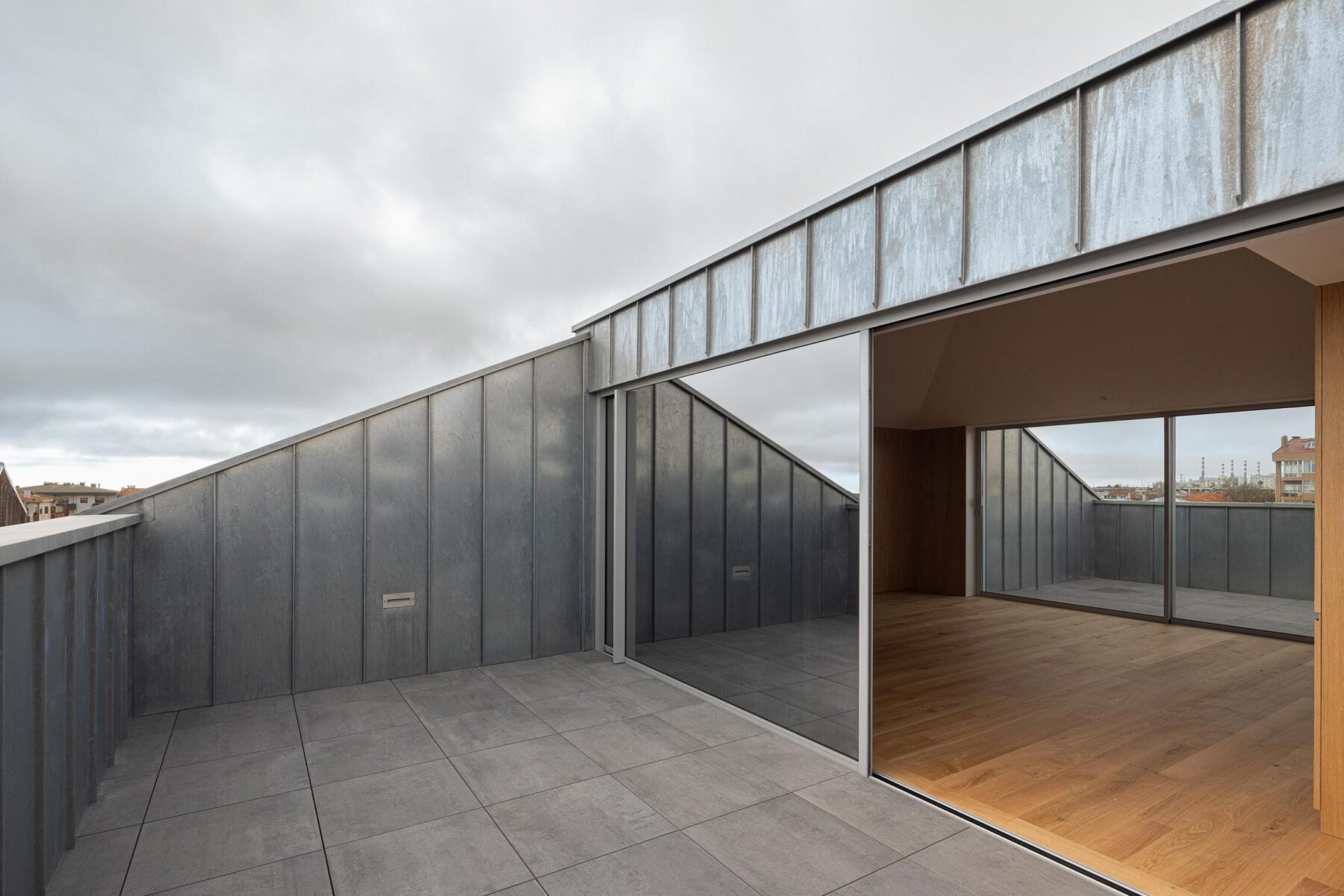 Terraço com pavimento cerâmico cinzento e revestimento metálico na cobertura inclinada, com um vão envidraçado para o interior. — EDIFÍCIO DE HABITAÇÃO MULTIFAMILIAR HINTZE RIBEIRO arquitectura HELDER DA ROCHA ARQUITECTOS fotografia Ivo Tavares