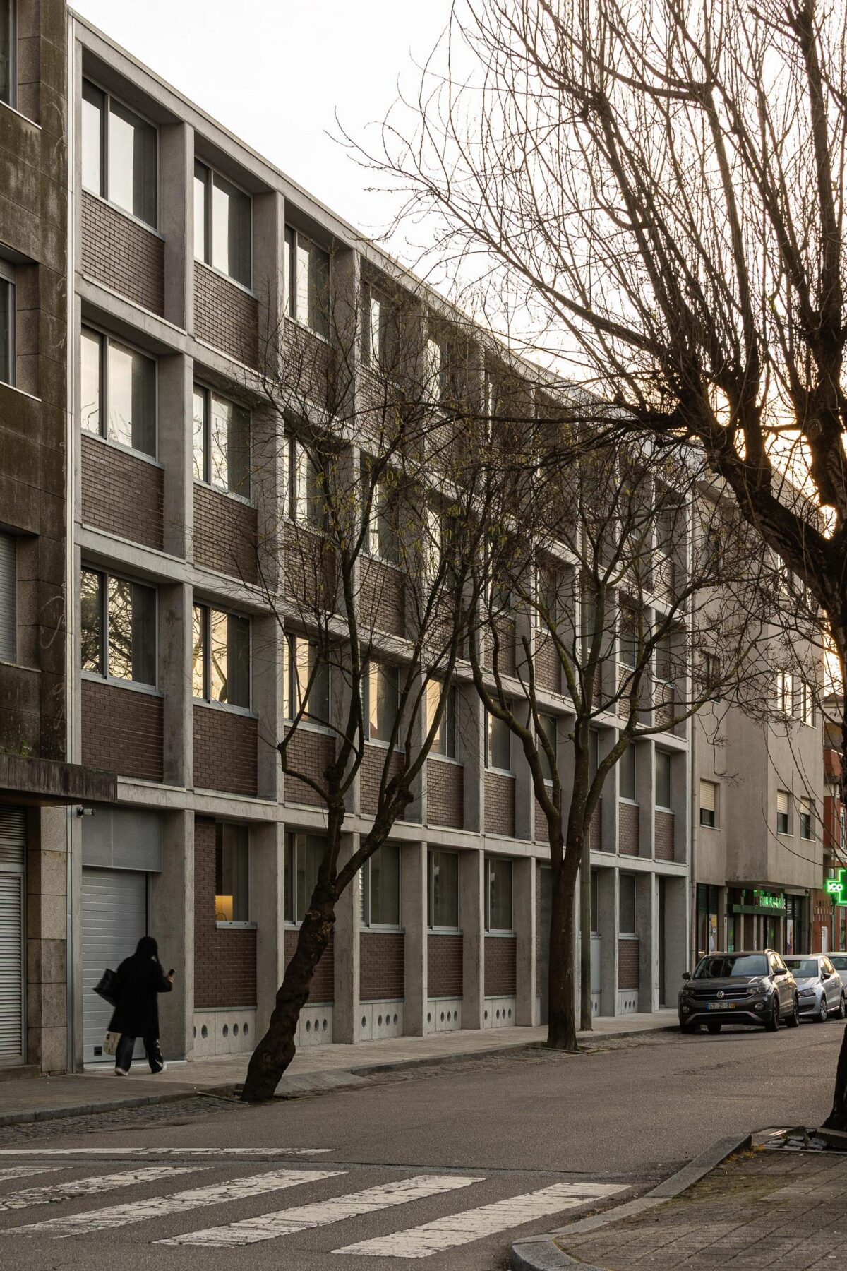 Edifício de habitação multifamiliar com fachada modular em betão aparente e tijolo, alinhado com a rua e a vegetação urbana. — EDIFÍCIO DE HABITAÇÃO MULTIFAMILIAR HINTZE RIBEIRO arquitectura HELDER DA ROCHA ARQUITECTOS fotografia Ivo Tavares