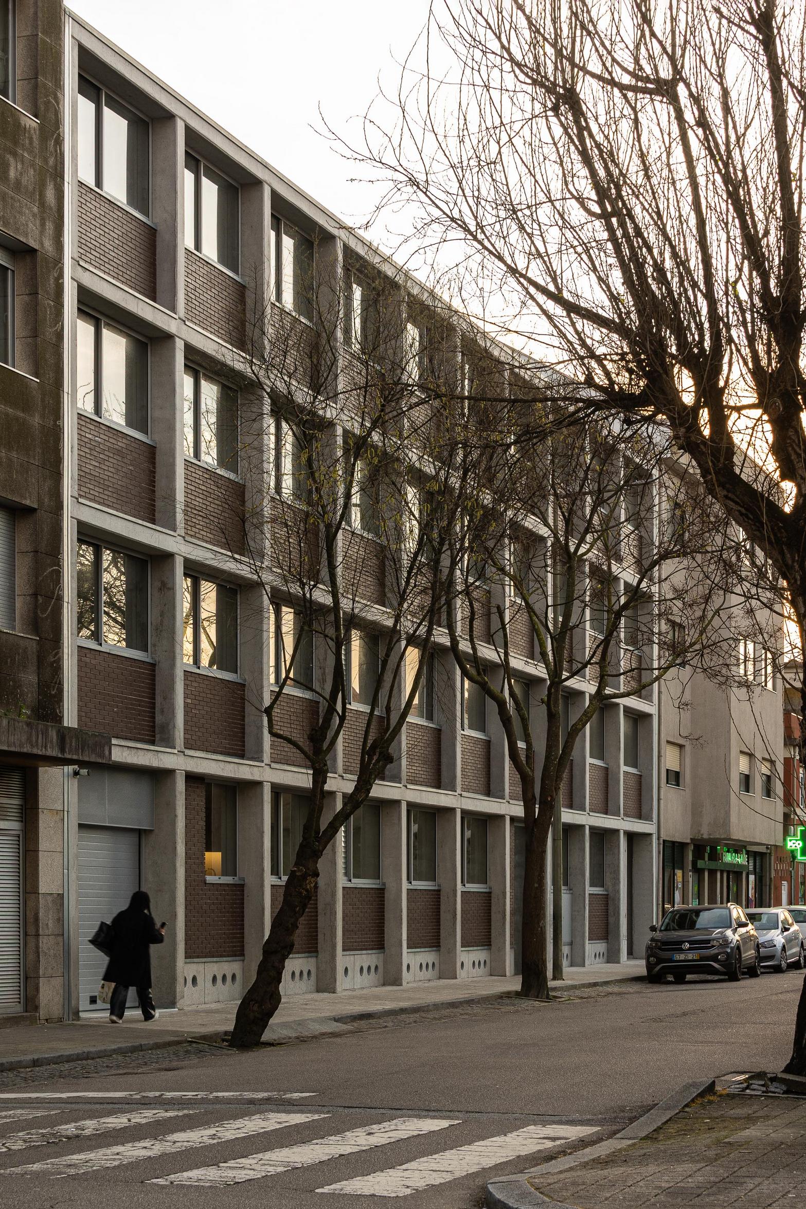Fachada urbana de betão aparente cinzento e tijolo maciço, com ritmo de vãos. Árvores sem folhas ladeiam o passeio — Edifício Multifamiliar Hintze Ribeiro arquitectura Helder da Rocha fotografia Ivo Tavares