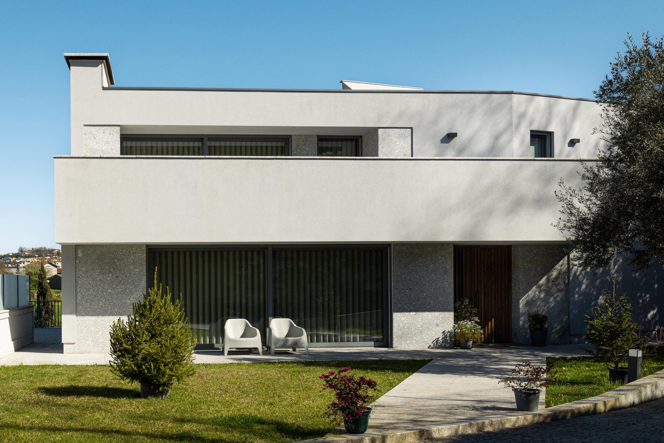 Fachada principal da moradia de dois pisos. Volumes brancos e embasamento em pedra. Grandes vãos envidraçados e jardim relvado — Duas Casas em Guimarães arquitectura Félix Arquitectos fotografia Ivo Tavares