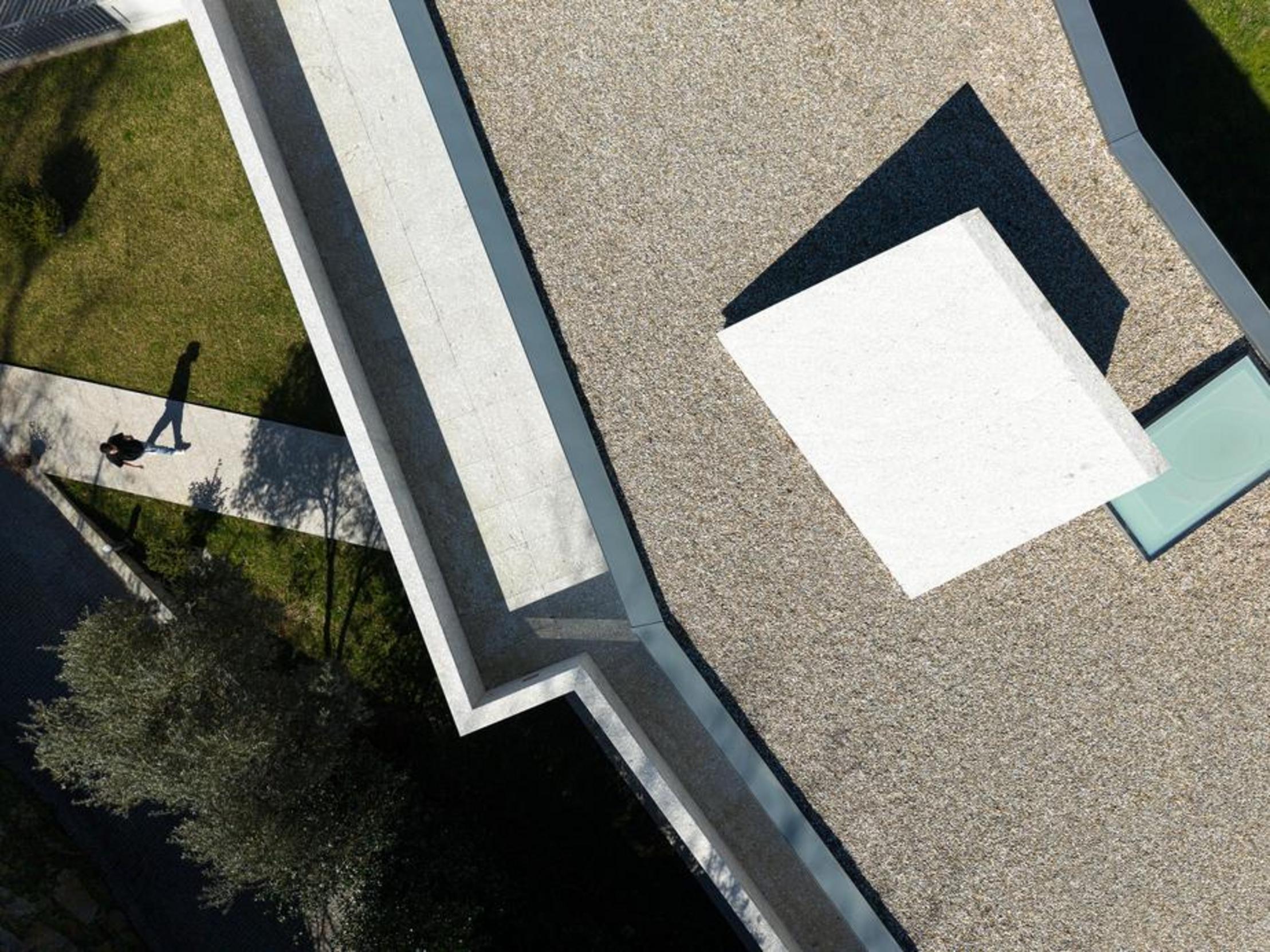 Vista aérea da cobertura. Pavimento em gravilha, caminhos em pedra clara. Volumes brancos e claraboia, com sombras projetadas no terreno — Duas Casas em Guimarães arquitectura Félix Arquitectos fotografia Ivo Tavares