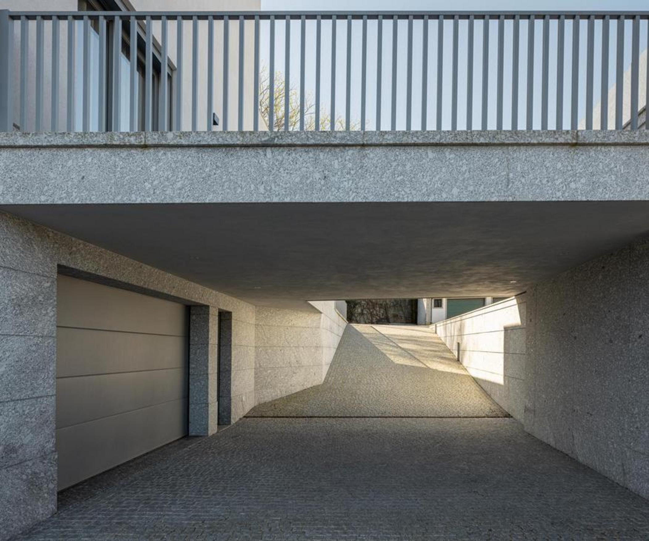 Garagem subterrânea com embasamento em pedra clara. Rampa de acesso em betão e vão de garagem metálico, iluminada por luz natural — Duas Casas em Guimarães arquitectura Félix Arquitectos fotografia Ivo Tavares
