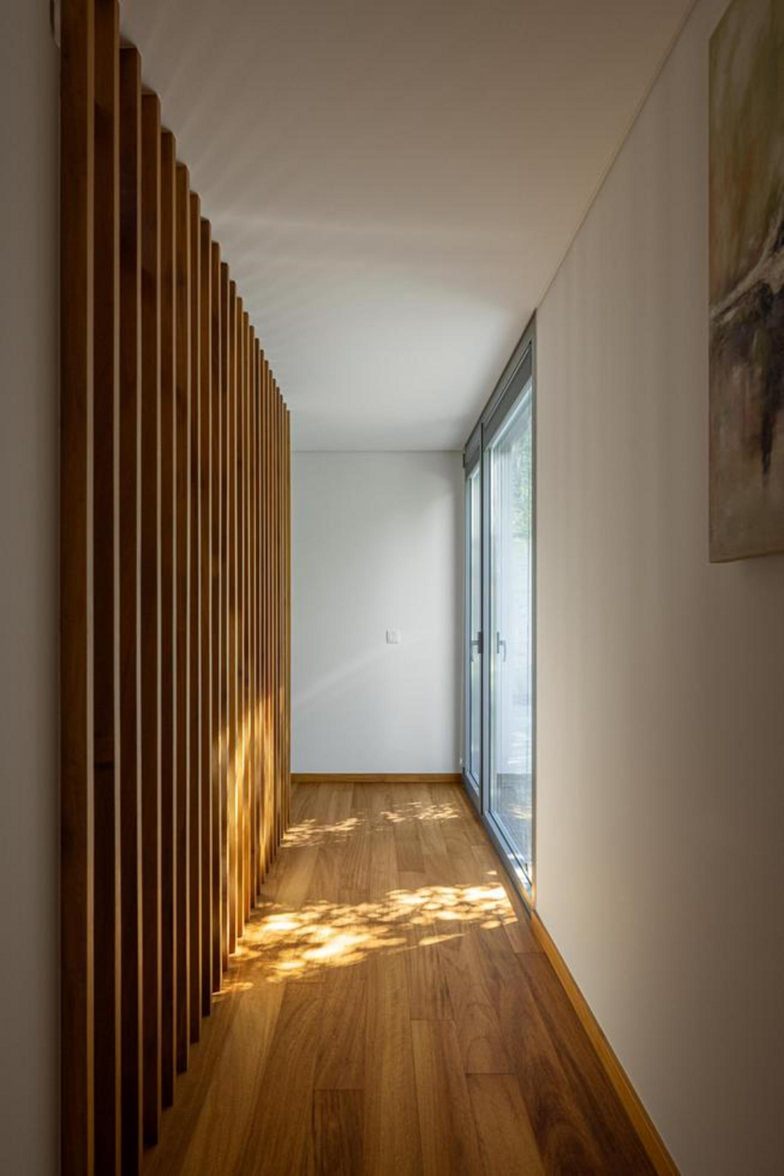 Corredor com pavimento em madeira e parede de ripas de madeira. Grande vão envidraçado permite entrada de luz natural e sombras — Duas Casas em Guimarães arquitectura Félix Arquitectos fotografia Ivo Tavares