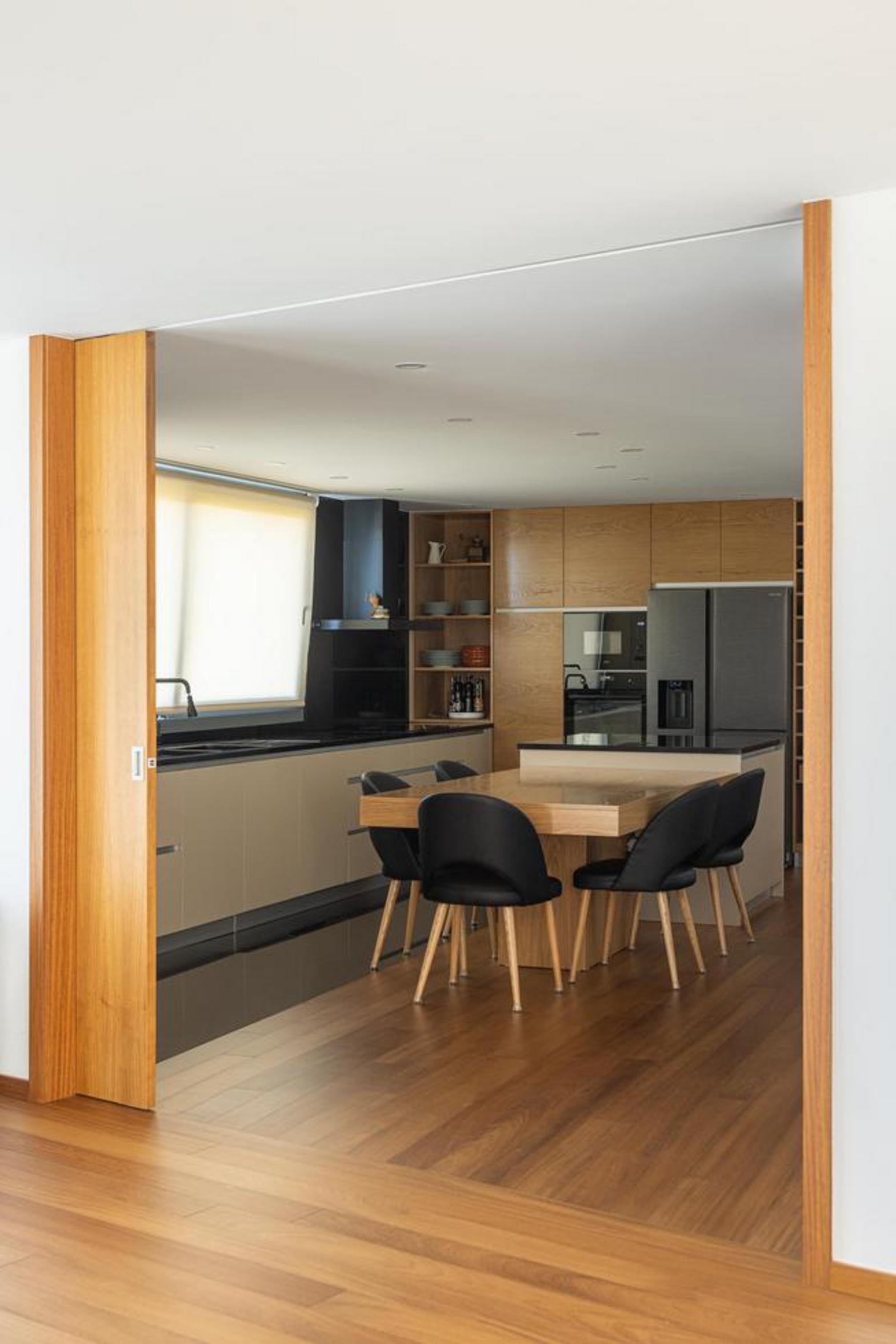 Cozinha com pavimento em madeira. Armários em madeira e cinzento. Mesa de jantar em madeira com cadeiras pretas, integrada no espaço — Duas Casas em Guimarães arquitectura Félix Arquitectos fotografia Ivo Tavares