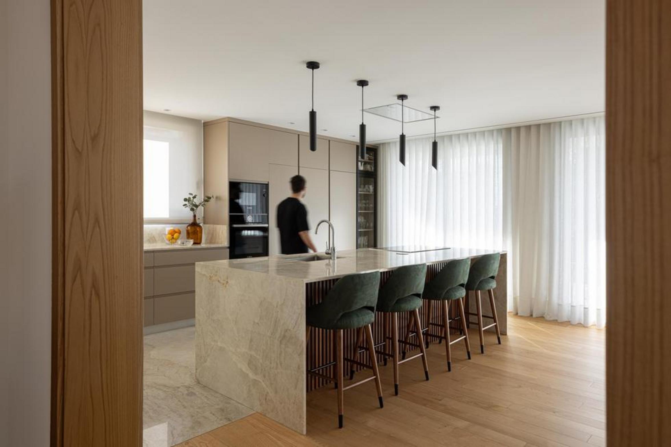Cozinha com ilha central em mármore bege e bancos estofados verdes. Armários neutros e pavimento em madeira e pedra — Duas Casas em Guimarães arquitectura Félix Arquitectos fotografia Ivo Tavares