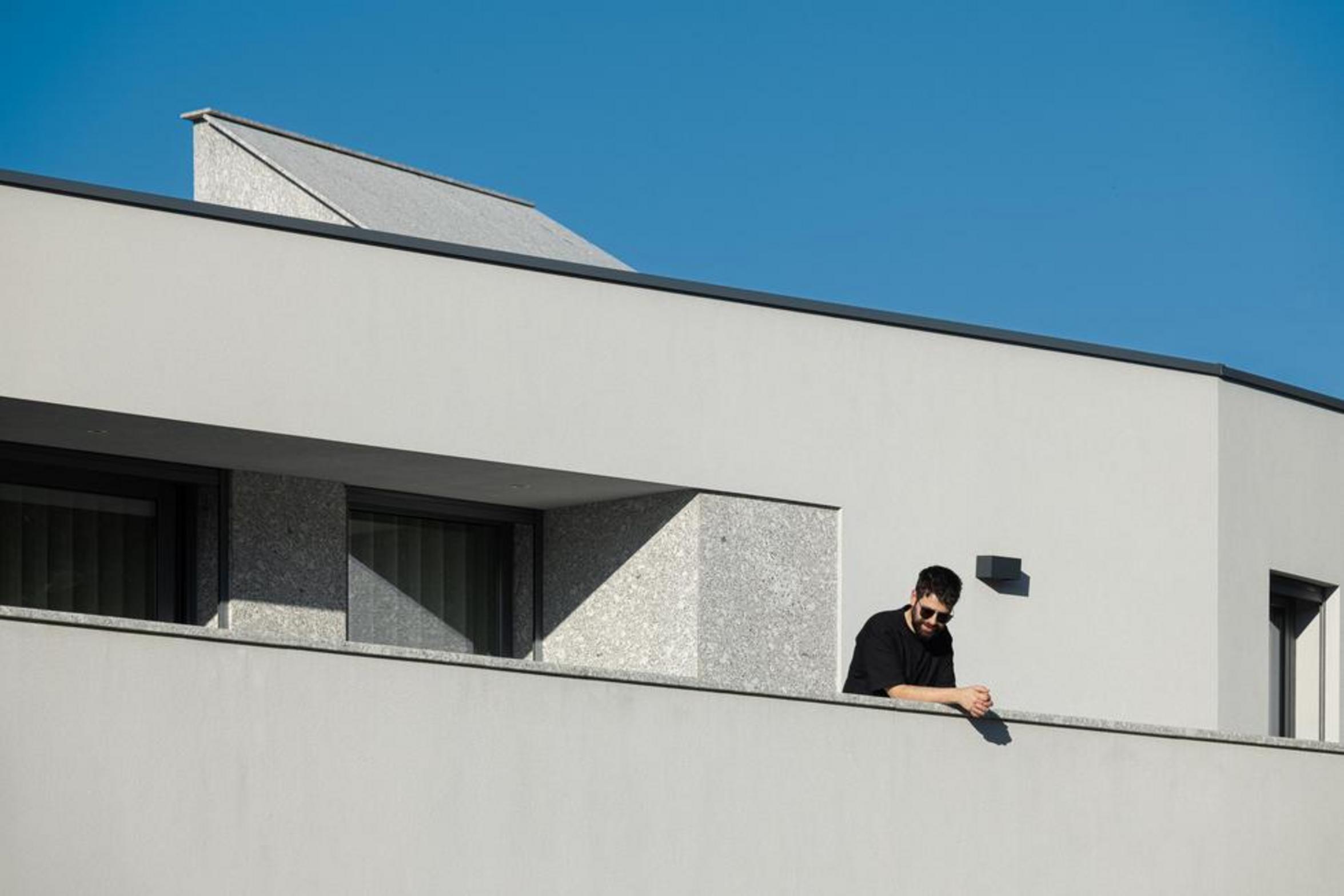 Varanda superior com fachada branca e apontamentos em pedra. Vãos retangulares e pessoa na varanda, sob um céu azul — Duas Casas em Guimarães arquitectura Félix Arquitectos fotografia Ivo Tavares