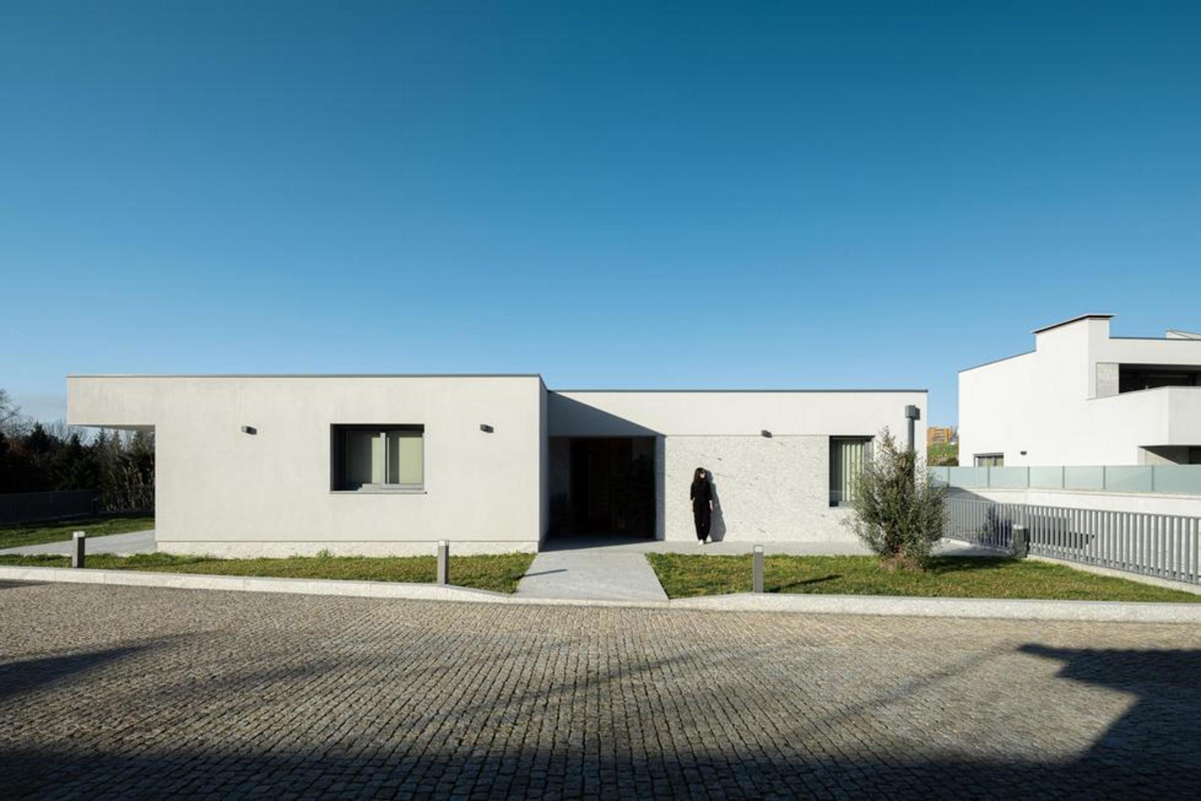 Fachada frontal da moradia térrea. Volume branco e embasamento em pedra. Entrada recuada e caminho em betão, ladeado por relva — Duas Casas em Guimarães arquitectura Félix Arquitectos fotografia Ivo Tavares