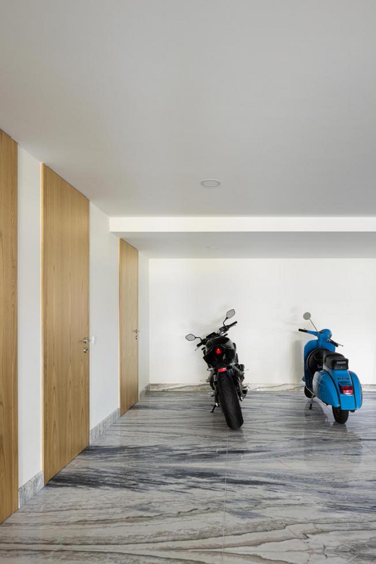 Garagem com pavimento em mármore de veios escuros. Paredes brancas e portas de madeira, com motos estacionadas no espaço — Duas Casas em Guimarães arquitectura Félix Arquitectos fotografia Ivo Tavares