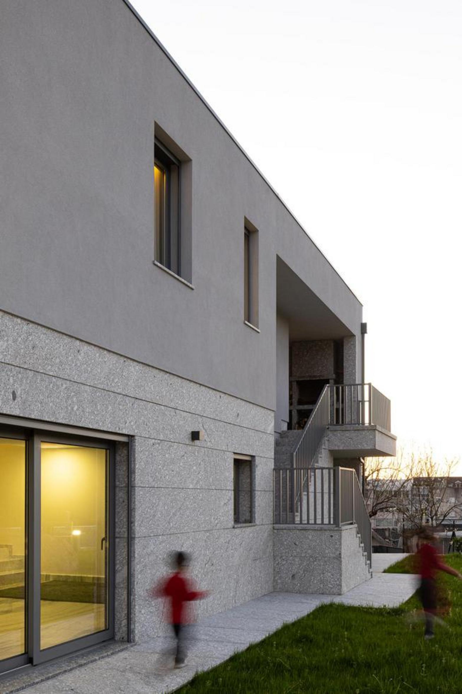 Fachada lateral com embasamento em pedra clara e volume superior cinzento. Escada exterior com guarda-corpos metálicos, integrando-se no terreno — Duas Casas em Guimarães arquitectura Félix Arquitectos fotografia Ivo Tavares