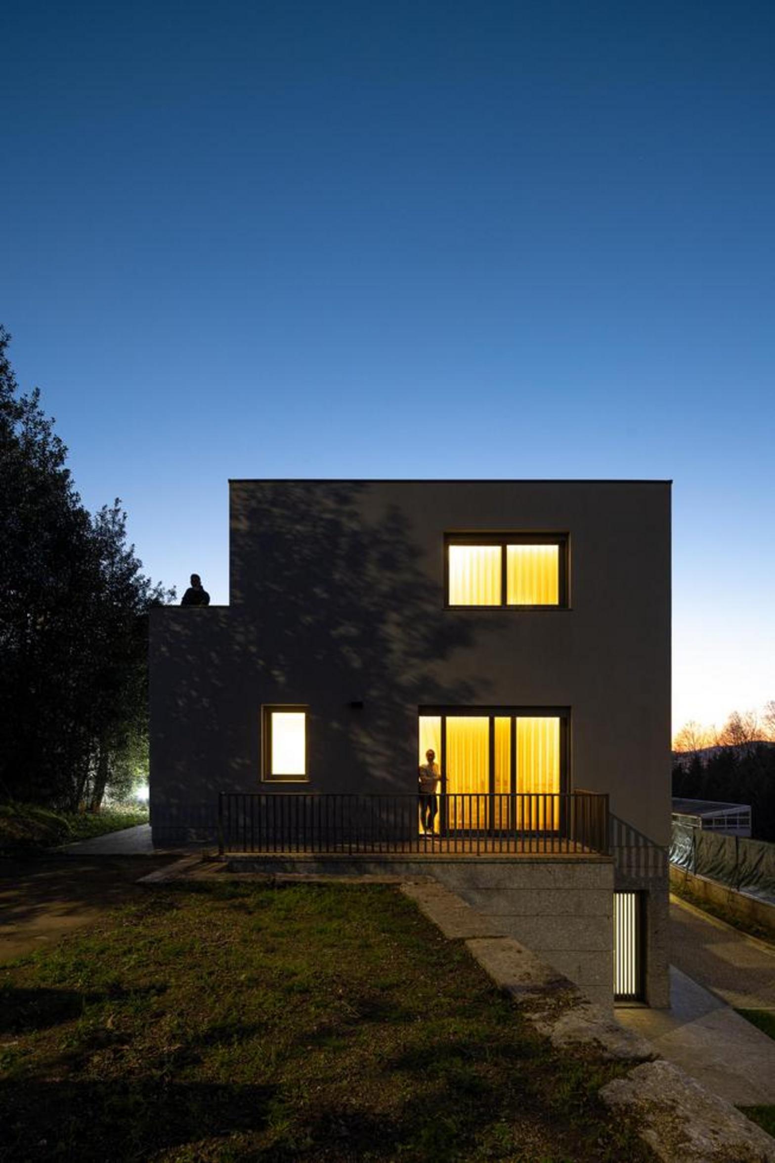 Fachada posterior da moradia ao anoitecer. Volumetria compacta em tom cinzento, com varanda e guarda-corpos metálicos. Iluminação interior quente — Duas Casas em Guimarães arquitectura Félix Arquitectos fotografia Ivo Tavares