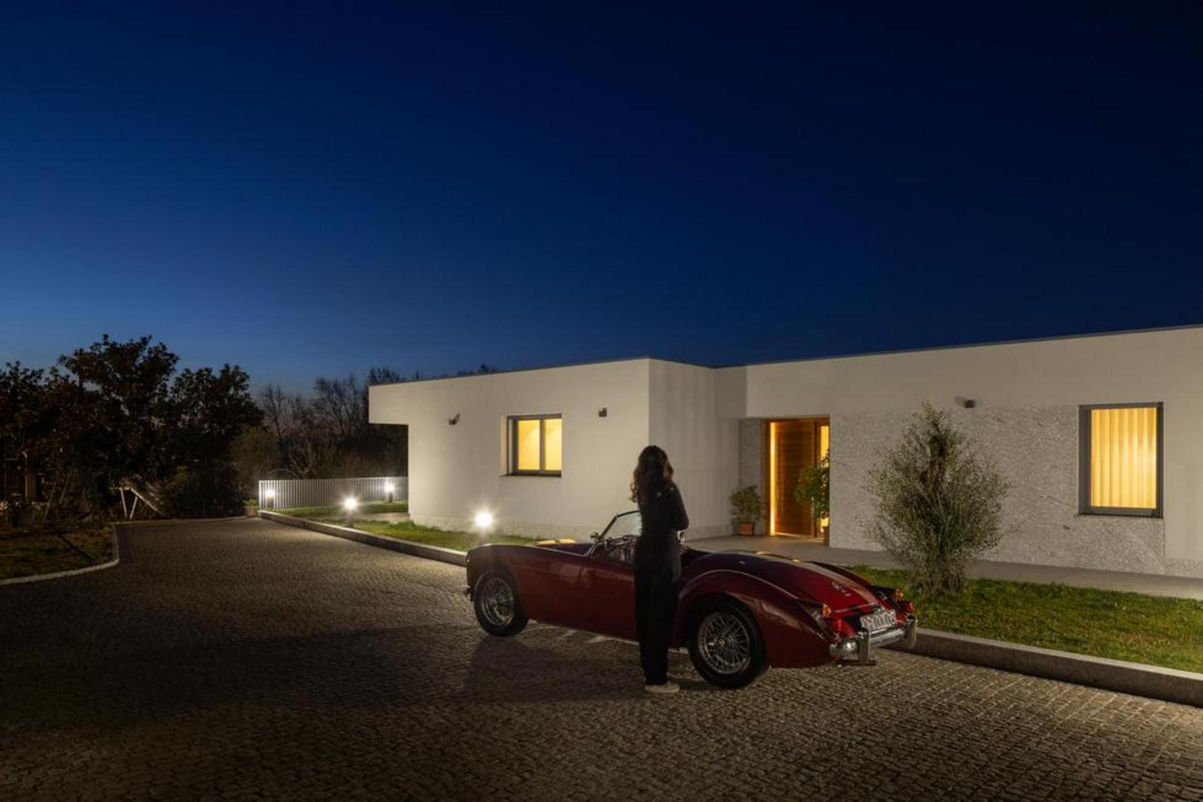 Fachada da moradia térrea ao anoitecer. Volume branco e embasamento em pedra. Entrada iluminada e caminho em calçada portuguesa — Duas Casas em Guimarães arquitectura Félix Arquitectos fotografia Ivo Tavares