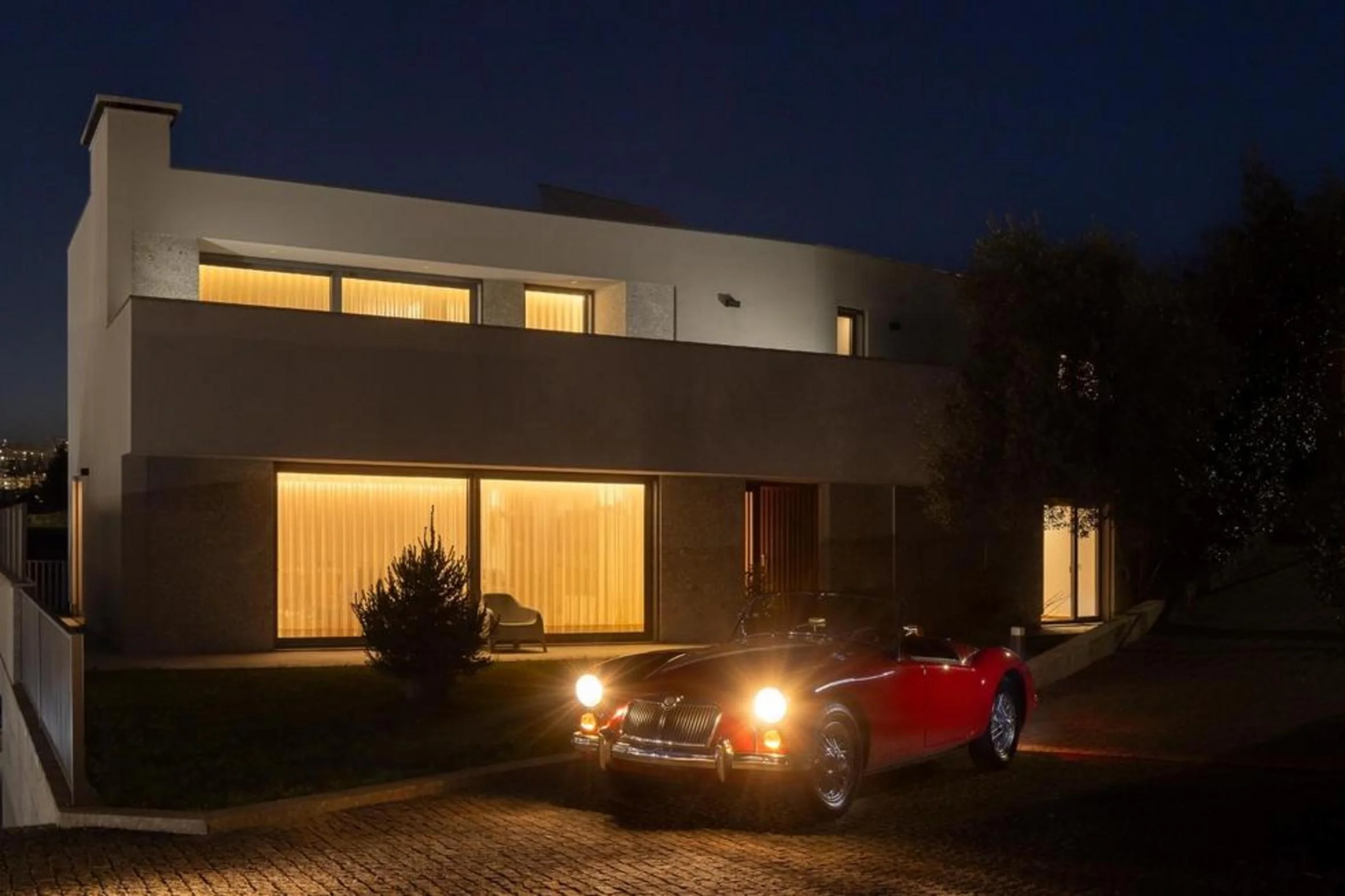 Fachada principal da moradia ao anoitecer. Volumes brancos e embasamento em pedra. Grandes vãos envidraçados com iluminação interior quente — Duas Casas em Guimarães arquitectura Félix Arquitectos fotografia Ivo Tavares