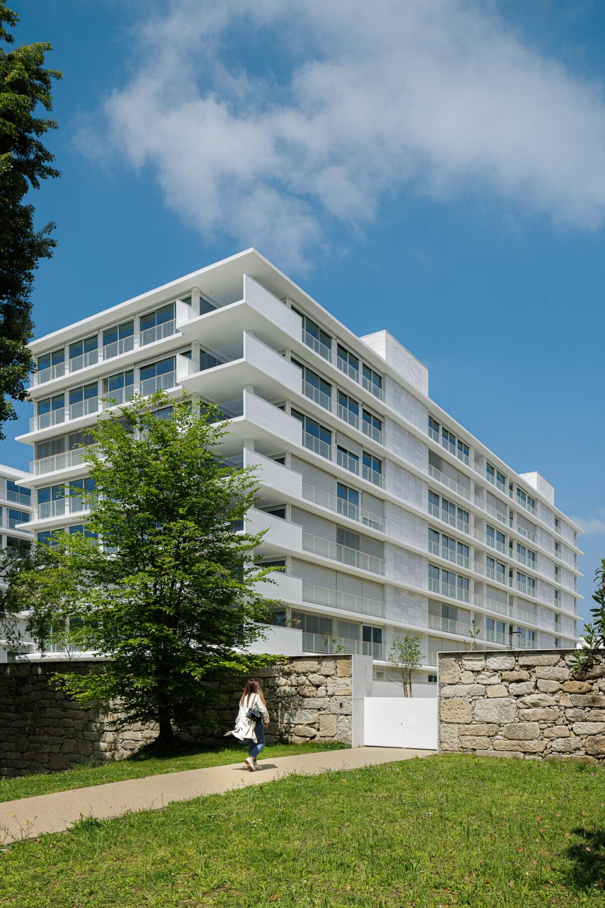 Fachada de edifício de apartamentos em reboco branco e vidro, com varandas em consola. Muro de pedra no contacto com o terreno — Edifício Natura arquitectura Visioarq Arquitectos fotografia Ivo Tavares