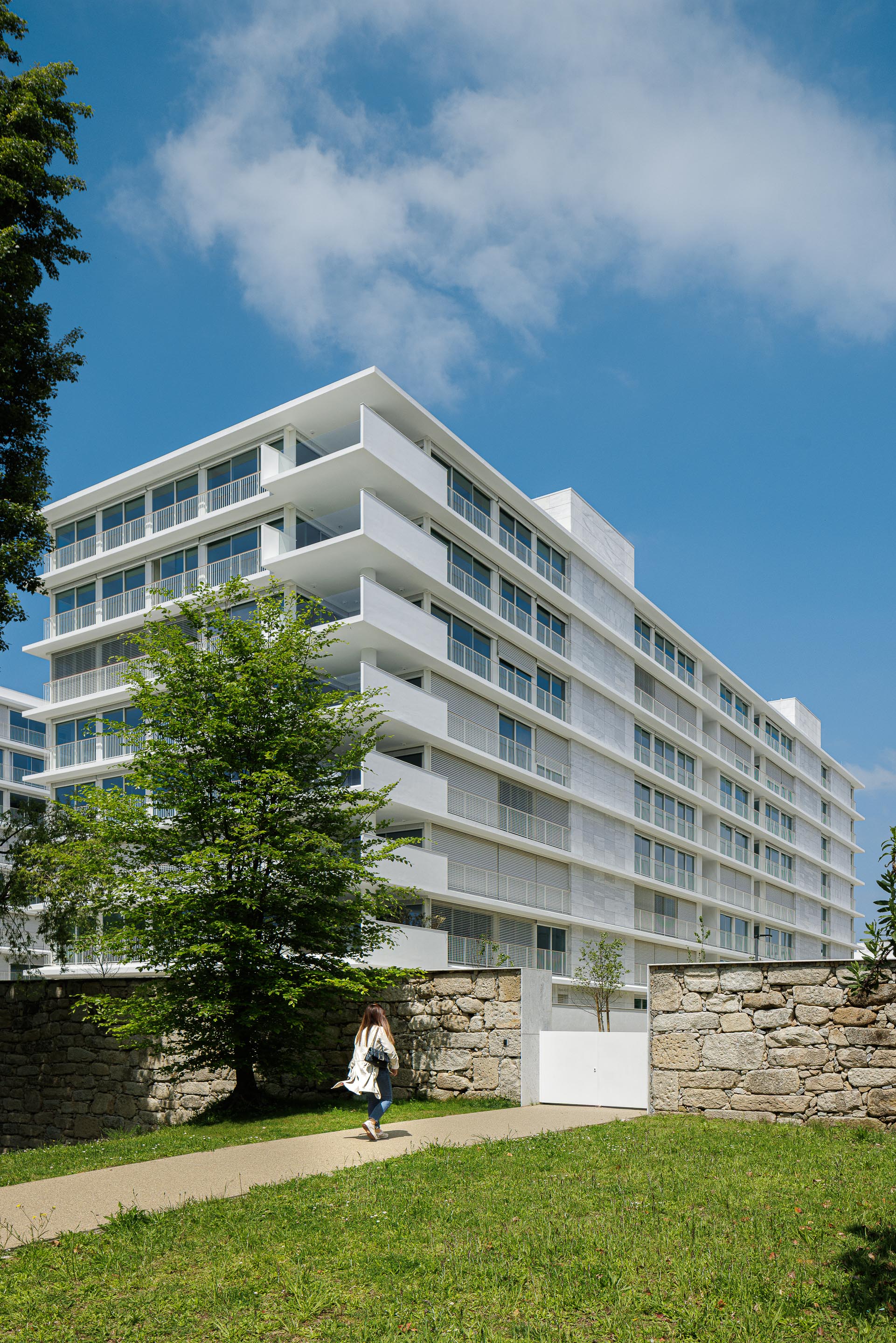 Fachada de edifício de apartamentos em reboco branco e vidro, com varandas em consola. Muro de pedra no contacto com o terreno — Edifício Natura arquitectura Visioarq Arquitectos fotografia Ivo Tavares