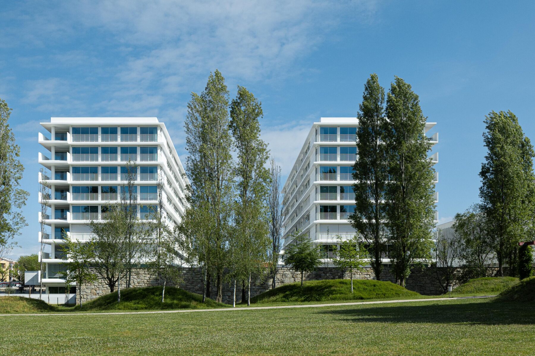 Duas fachadas de edifícios de apartamentos brancos, com grandes vãos envidraçados, inseridos num parque verde com muro de pedra — Edifício Natura arquitectura Visioarq Arquitectos fotografia Ivo Tavares