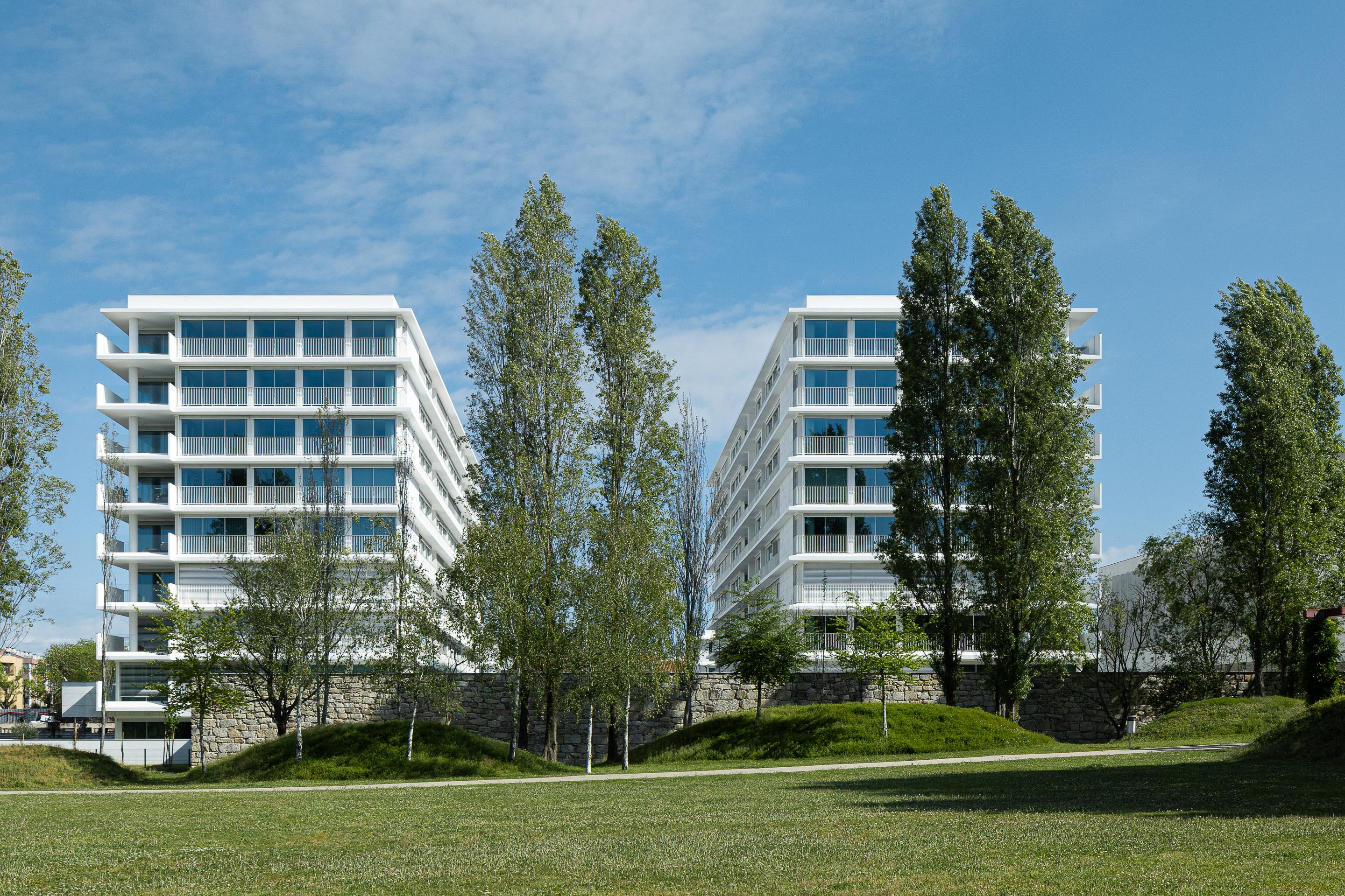 Duas fachadas de edifícios de apartamentos brancos, com grandes vãos envidraçados, inseridos num parque verde com muro de pedra — Edifício Natura arquitectura Visioarq Arquitectos fotografia Ivo Tavares
