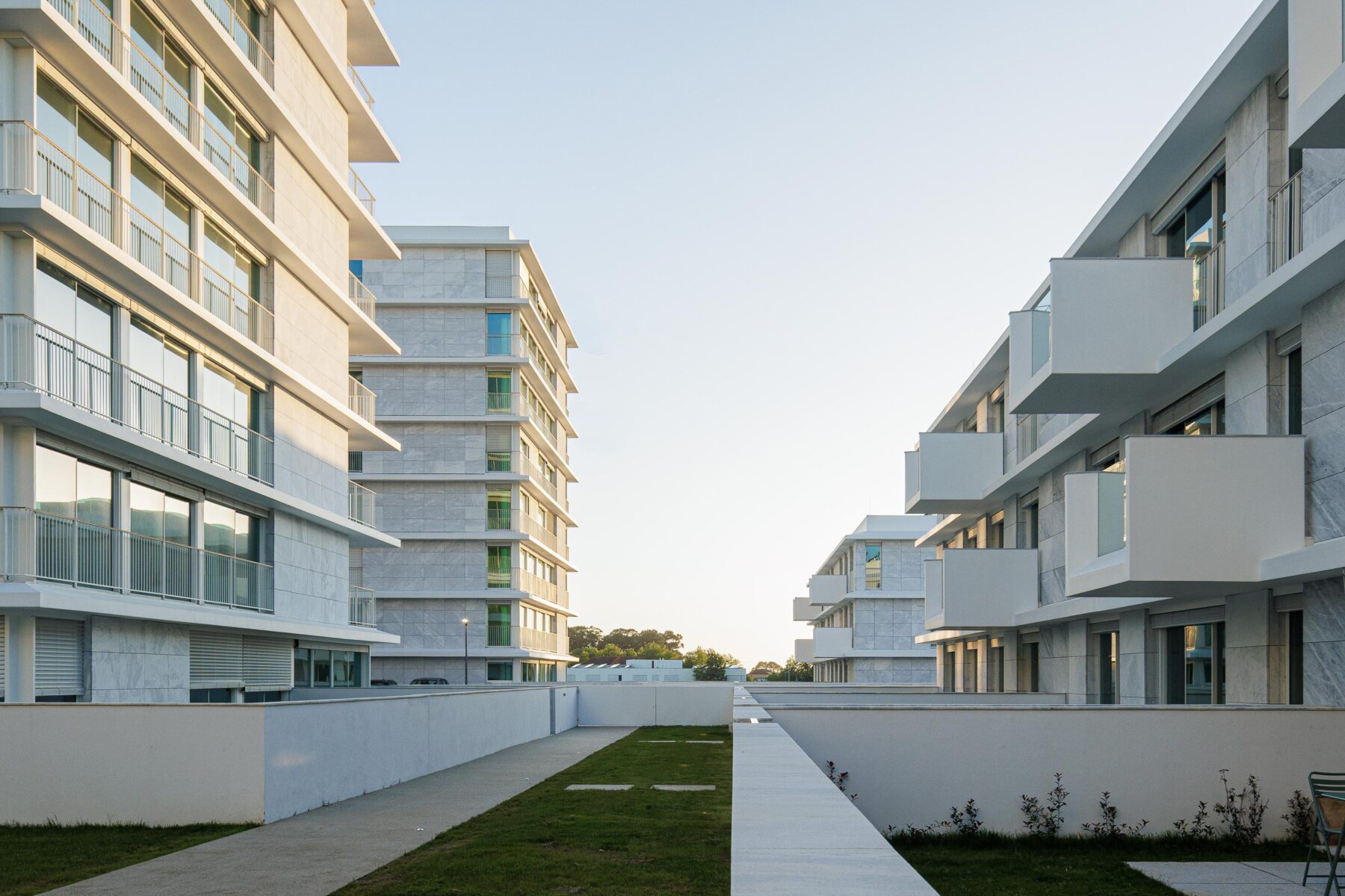Pátio ajardinado com relva e paredes em reboco branco, ladeado por edifícios com fachadas em mármore branco — Edifício Natura arquitectura Visioarq Arquitectos fotografia Ivo Tavares