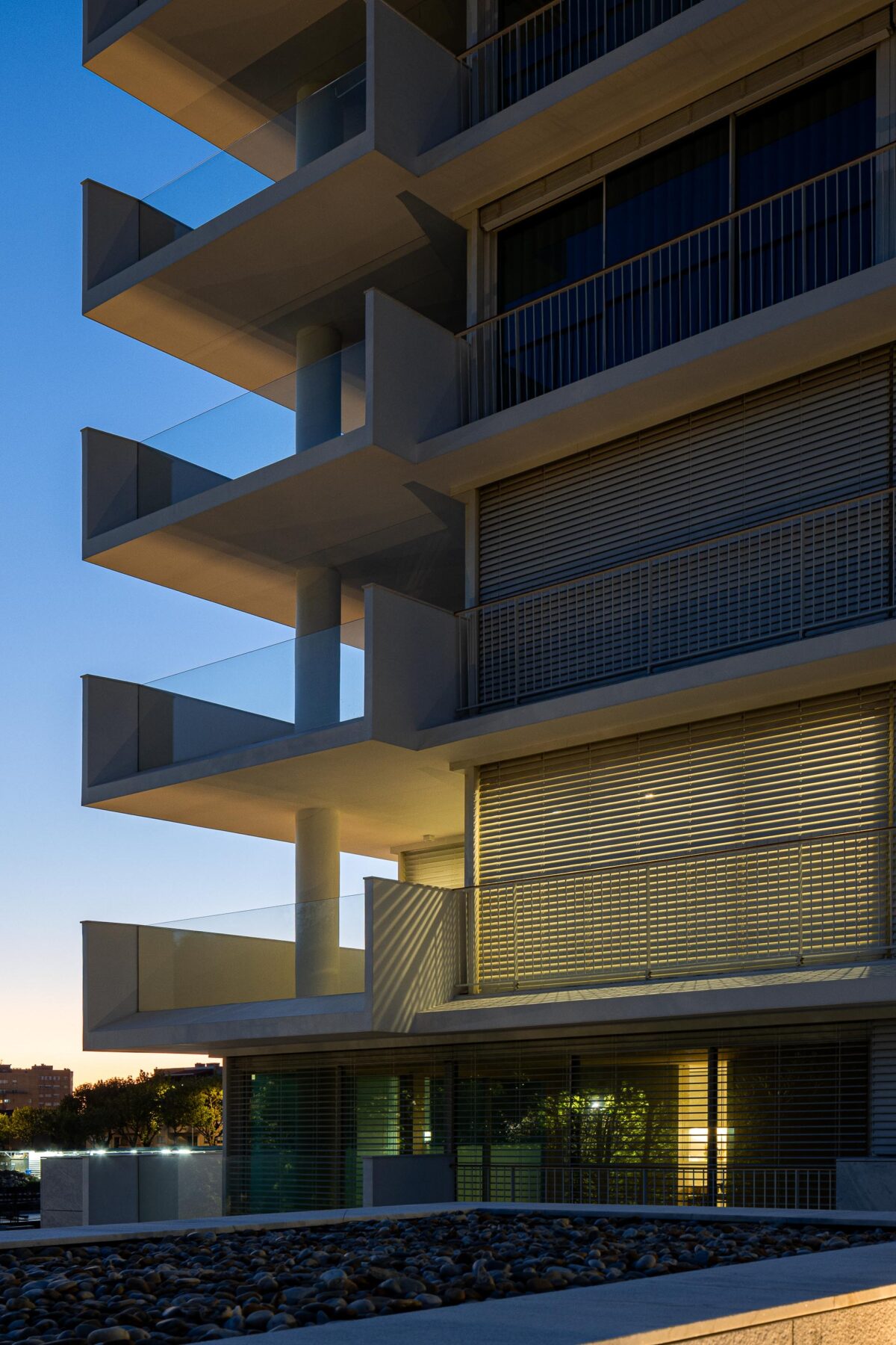 Fachada de edifício ao entardecer, com varandas em consola e pilares cilíndricos. Grandes vãos envidraçados com estores — Edifício Natura arquitectura Visioarq Arquitectos fotografia Ivo Tavares