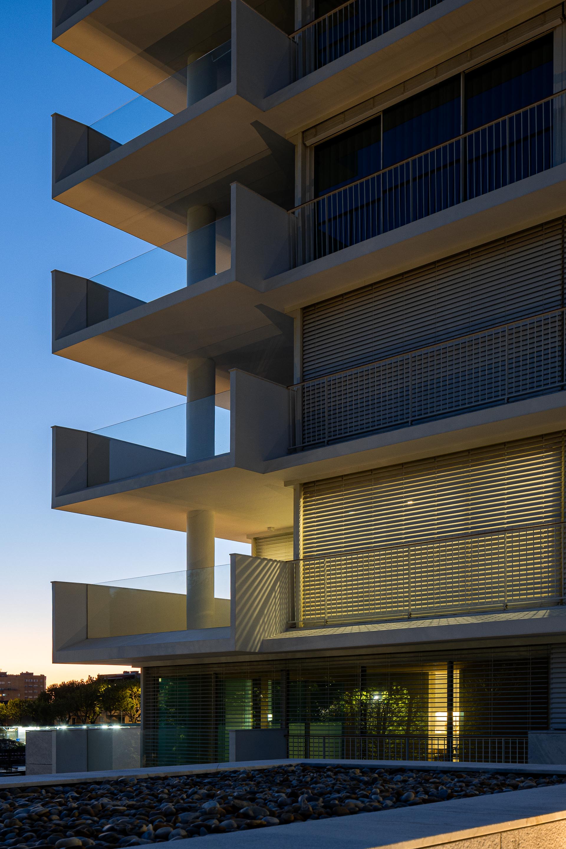 Fachada de edifício ao entardecer, com varandas em consola e pilares cilíndricos. Grandes vãos envidraçados com estores — Edifício Natura arquitectura Visioarq Arquitectos fotografia Ivo Tavares