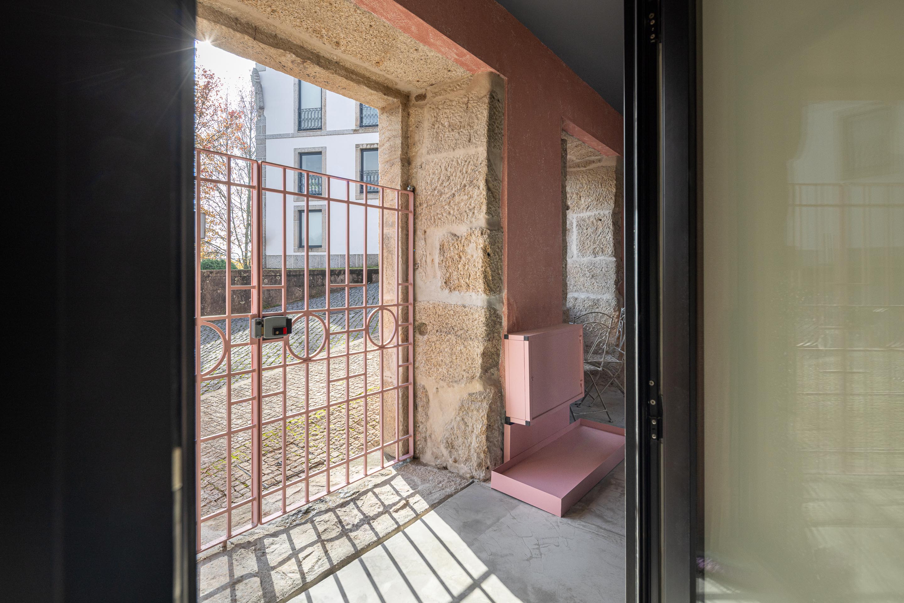 Entrada com portão metálico rosa, pavimento em betão e parede de pedra granítica exposta — Boutique Apartments arquitectura HPA fotografia Ivo Tavares