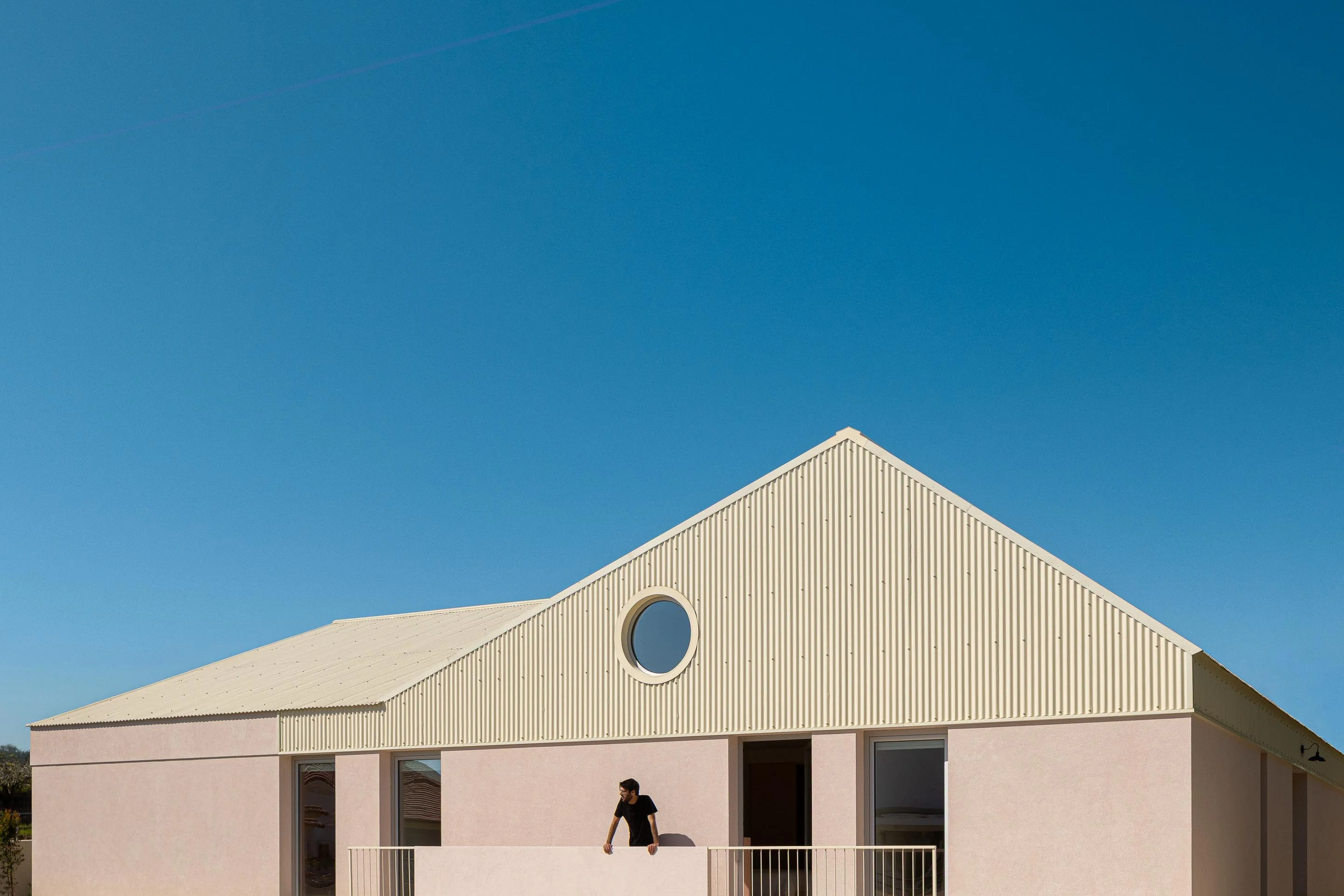 Moradia com fachada em estuque rosa e cobertura em chapa metálica bege, com pessoa na varanda — Casa do Alpendre arquitectura Vasco Burnay Arquitectura fotografia Ivo Tavares