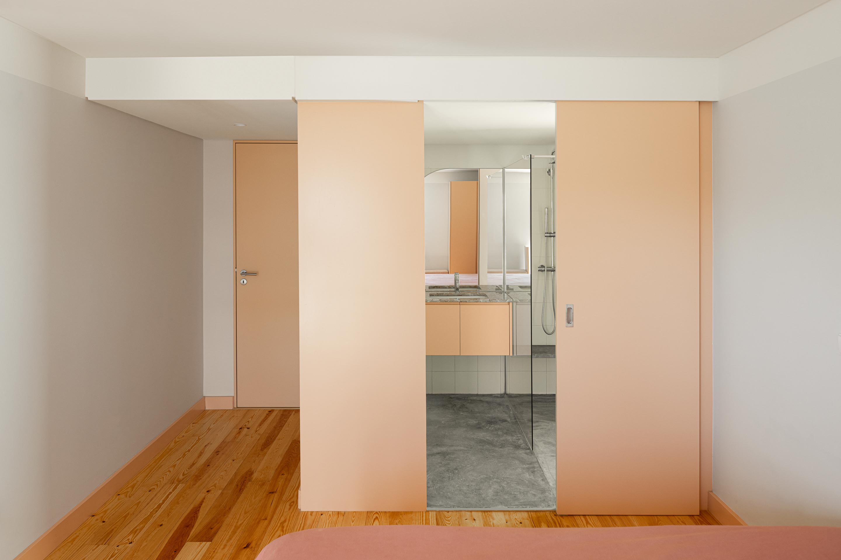 Quarto com pavimento em madeira e porta de correr pêssego, revelando casa de banho adjacente — Casa do Alpendre arquitectura Vasco Burnay Arquitectura fotografia Ivo Tavares
